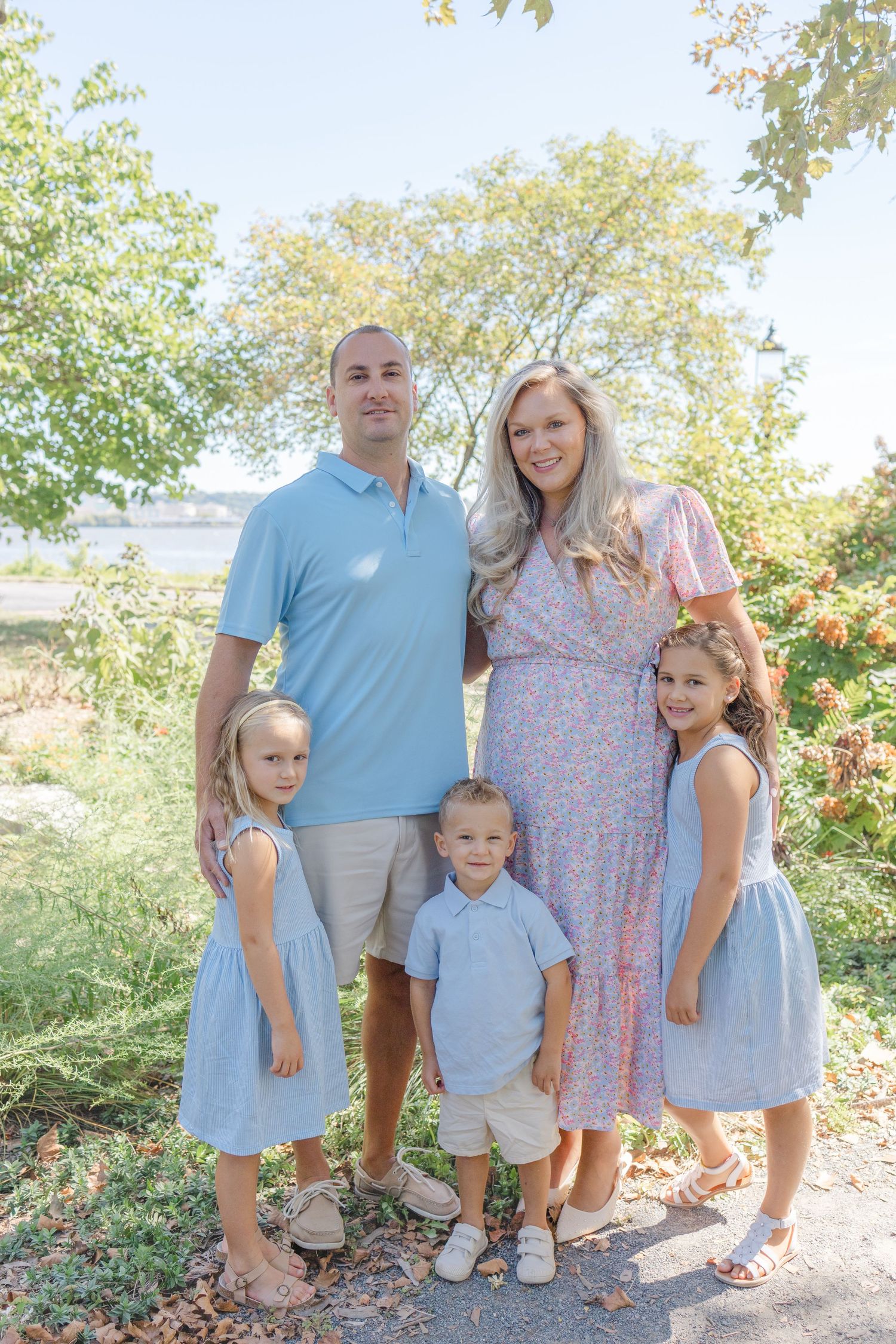 Family of five posing outdoors in coordinating blue outfits, with two girls, a toddler boy, mom, and dad smiling.