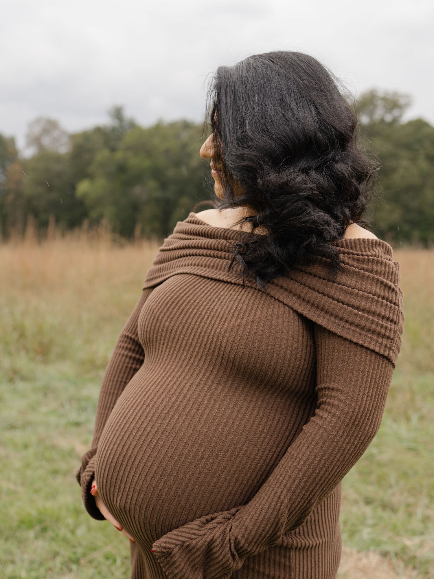 Pregnant woman in brown ribbed off-shoulder dress standing in a field, cradling her baby bump outdoors.