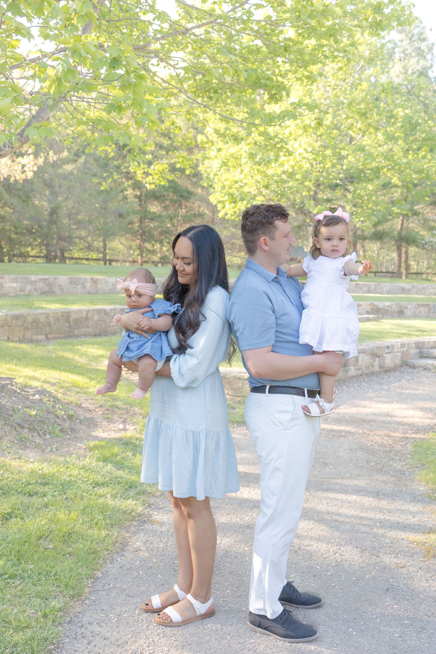 A young couple stands back-to-back outdoors, each holding a baby dressed in white and blue, surrounded by lush green trees.