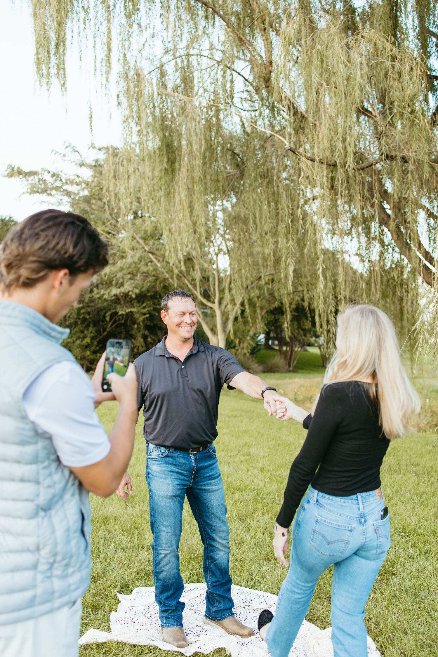 A man and woman hold hands outdoors on a blanket while another person photographs them near a willow tree.