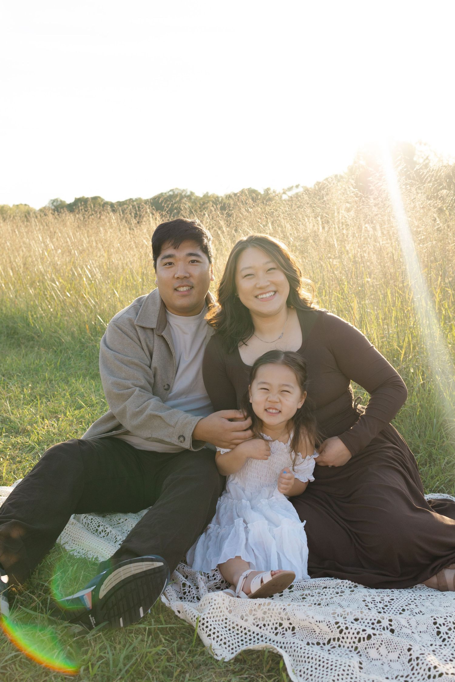 Family of three smiling outdoors on a blanket in a sunlit grassy field, parents with young daughter in white dress.