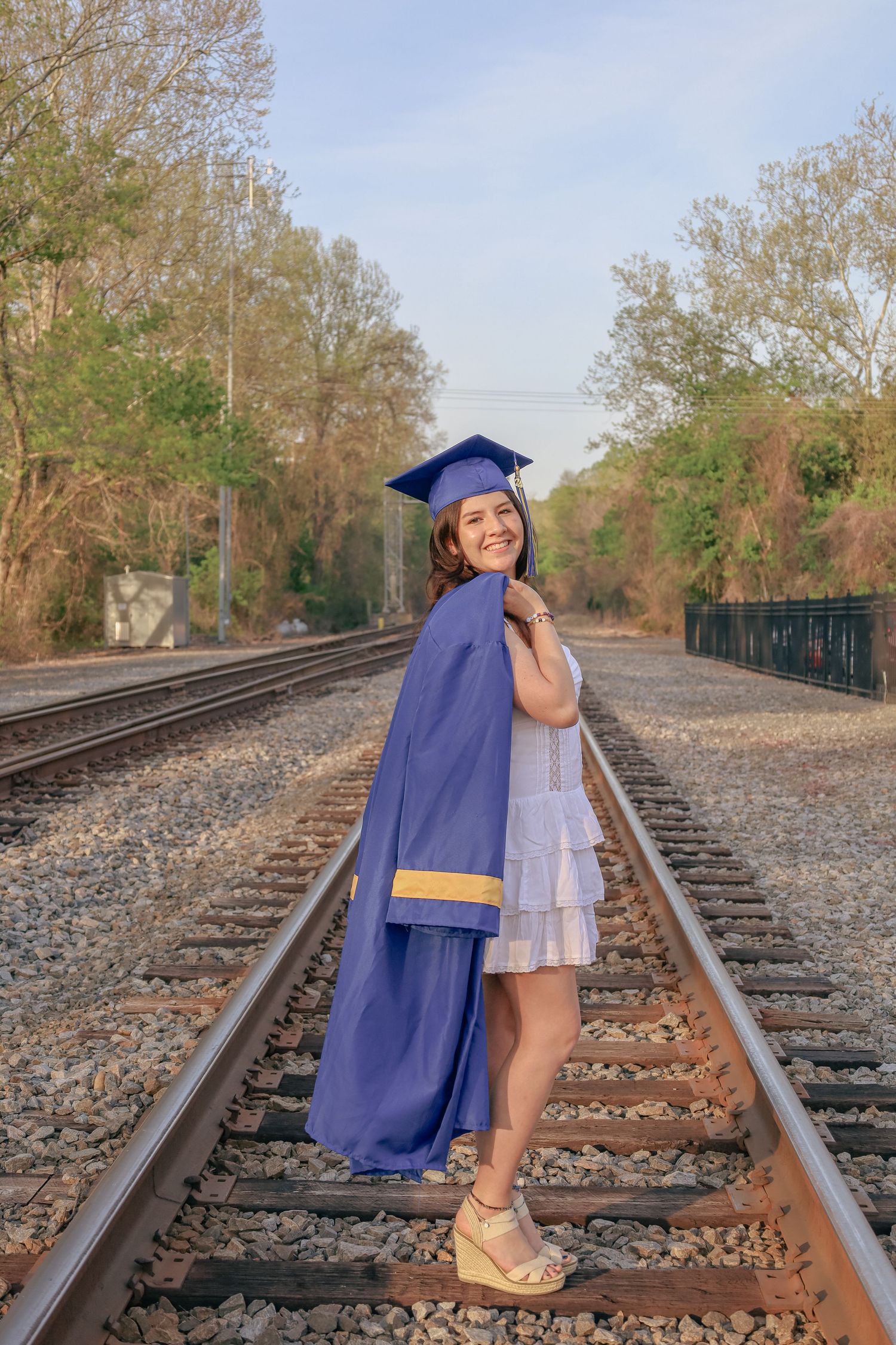 Graduate in blue cap and gown poses on train tracks during sunset.