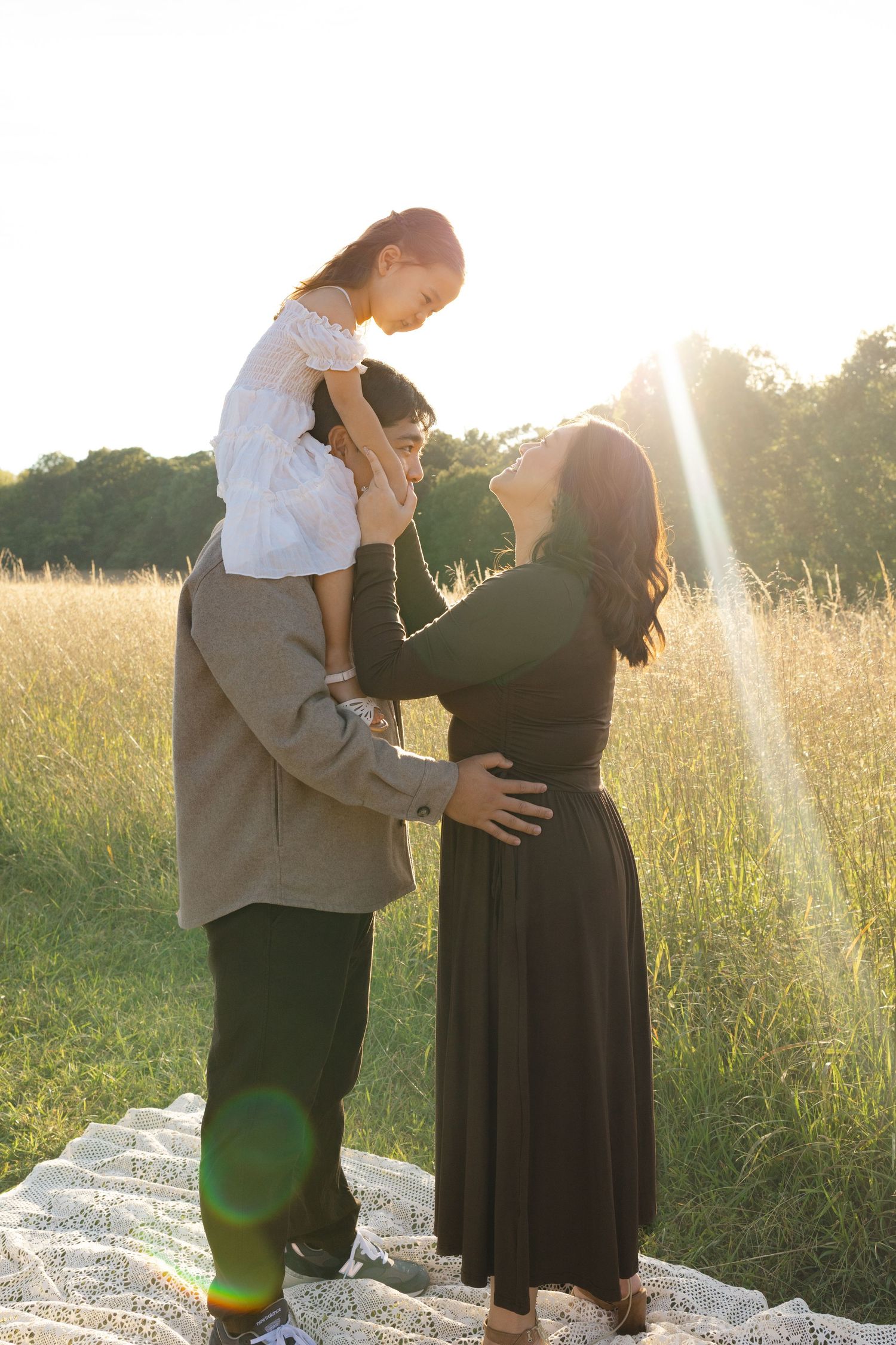 Family maternity photo outdoors at golden hour, father holding child on shoulders while pregnant mother looks up lovingly.