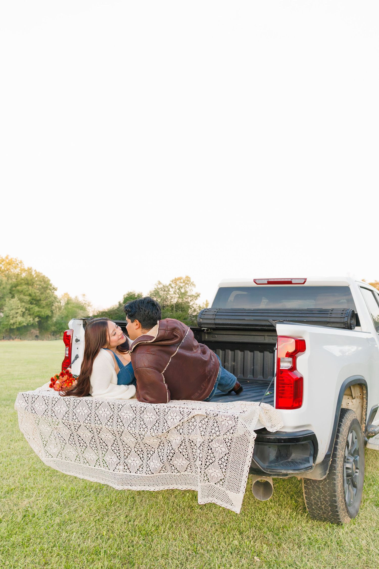 Couple sharing a romantic kiss on the tailgate of a white pickup truck, covered with a lace blanket in a green field.
