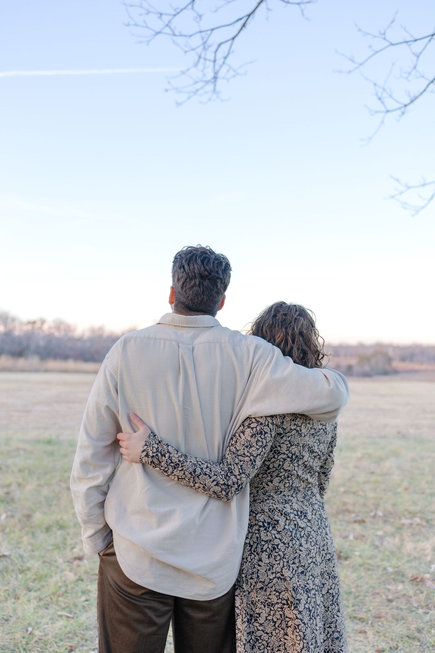 Couple embracing from behind, standing in an open field during autumn, wearing casual earth-toned clothing.