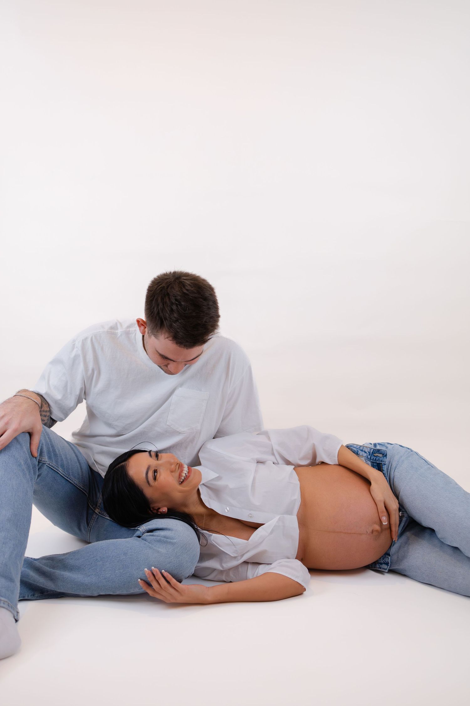 Couple expecting a baby, woman lying down showing bump, man sitting beside her smiling warmly.