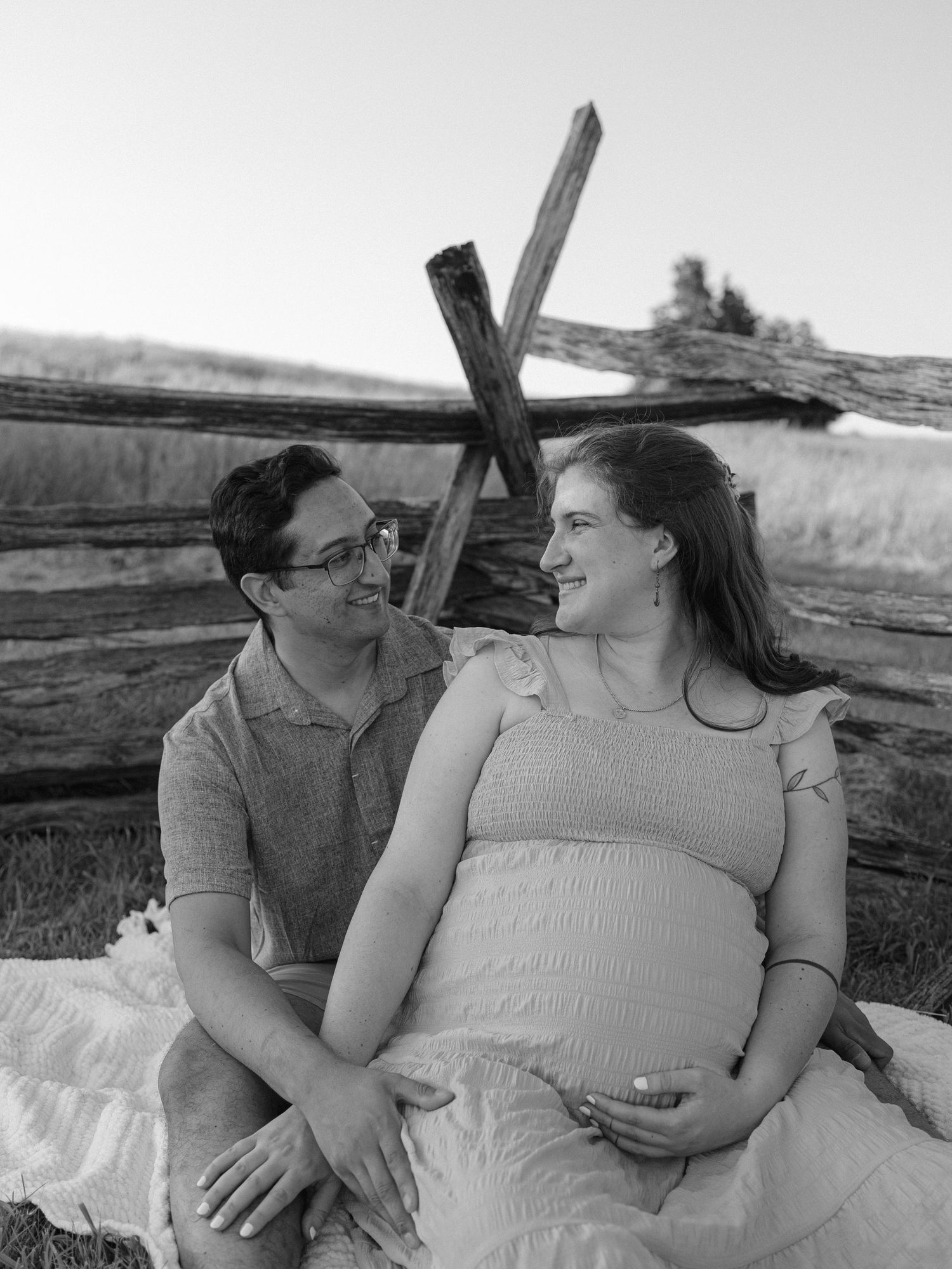 Black and white maternity photo of a couple sitting together outdoors near a rustic wooden fence.