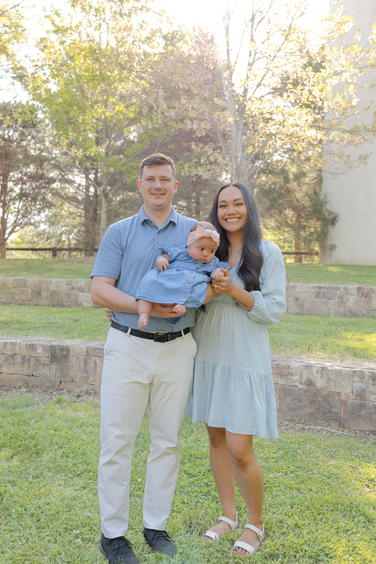 A smiling couple stands outdoors on green grass holding a baby dressed in blue, surrounded by trees in soft natural light.