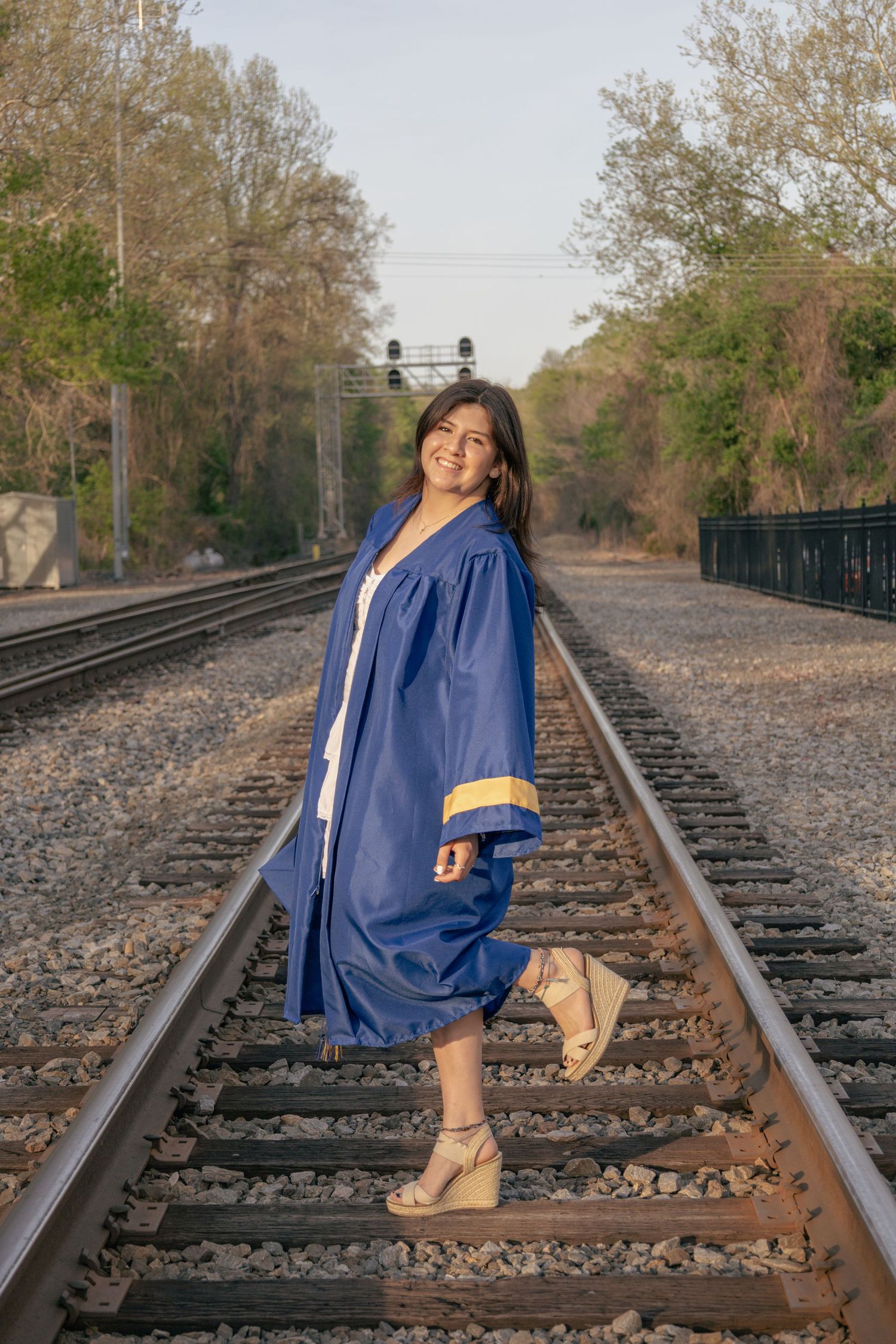 Graduate in blue cap and gown poses playfully on train tracks during golden hour.