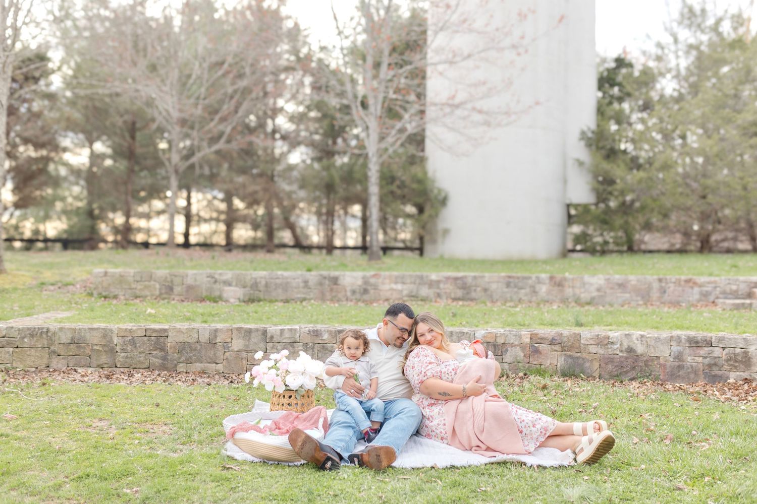 Expecting family sits on pink blanket in park, parents with toddler, maternity photo shoot outdoors.