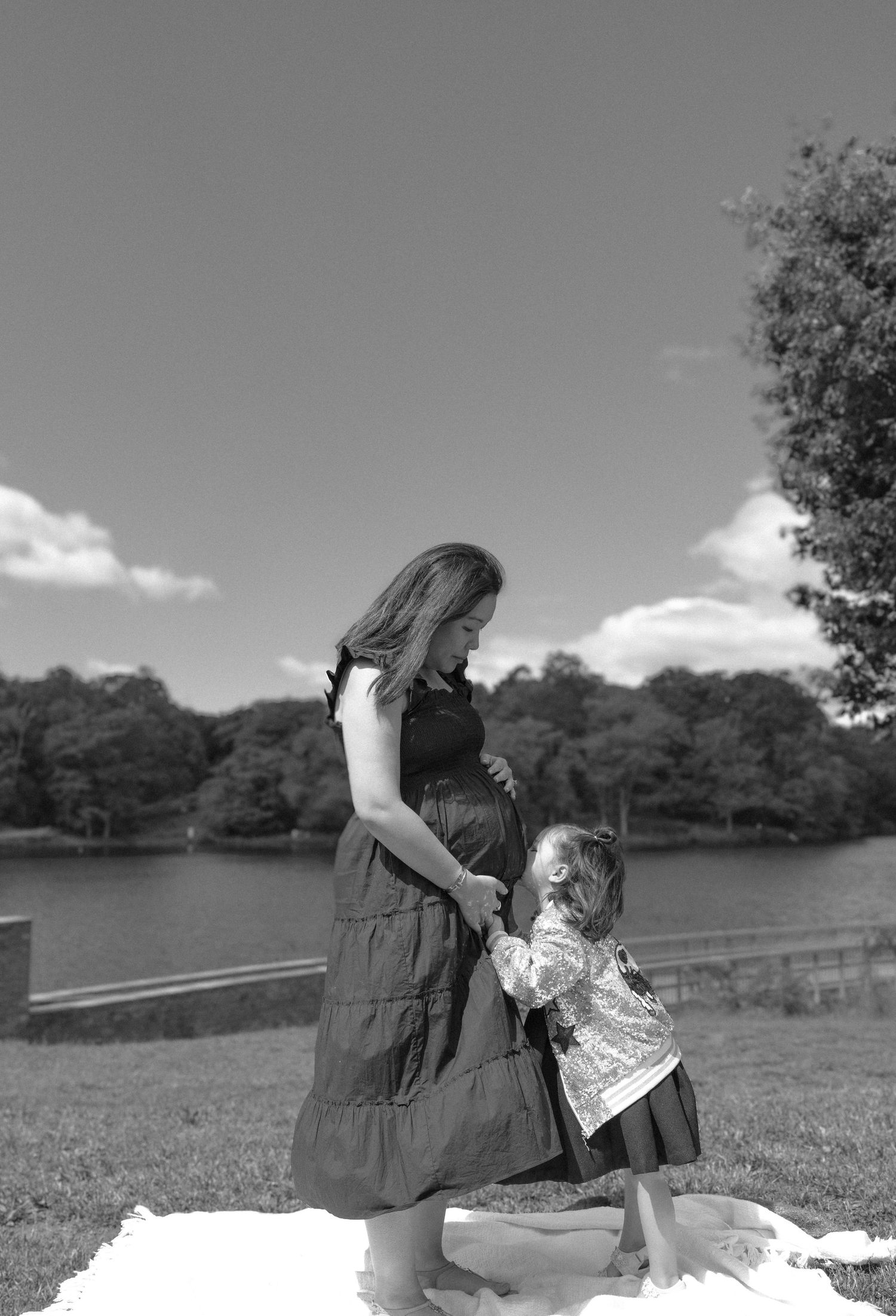 Black and white photo of pregnant woman holding hands with young daughter outdoors near a lake with trees in background.