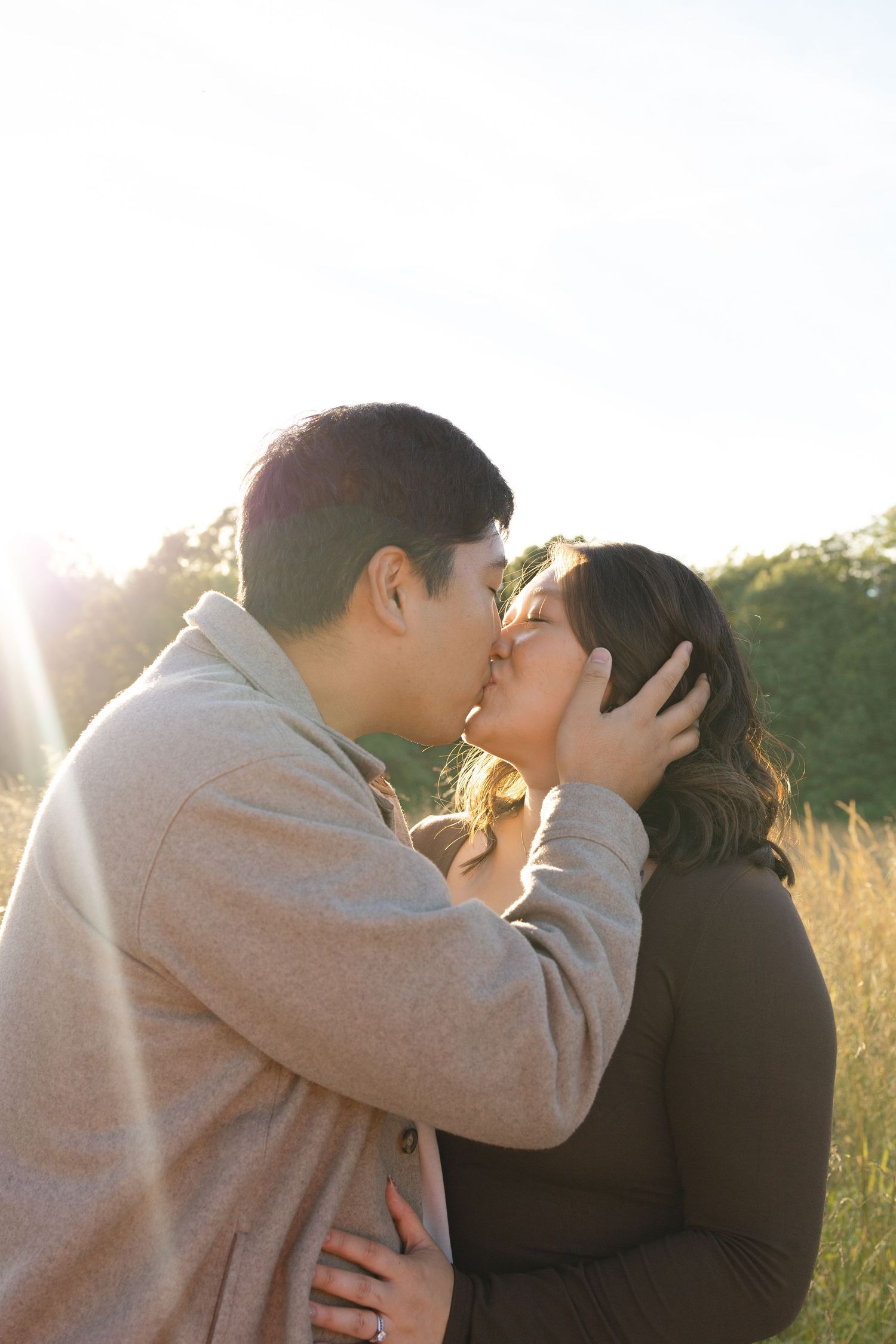 A couple shares a romantic kiss in a sunlit field, the man tenderly holding the woman's face in golden hour light.