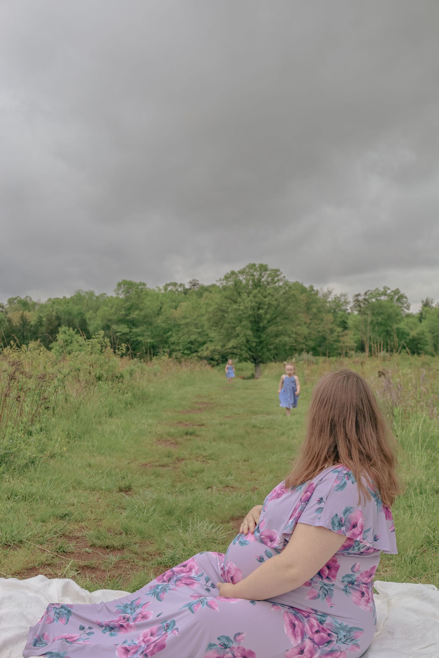 Pregnant woman in floral dress sits on blanket in green field under cloudy sky, with figures walking in distance.