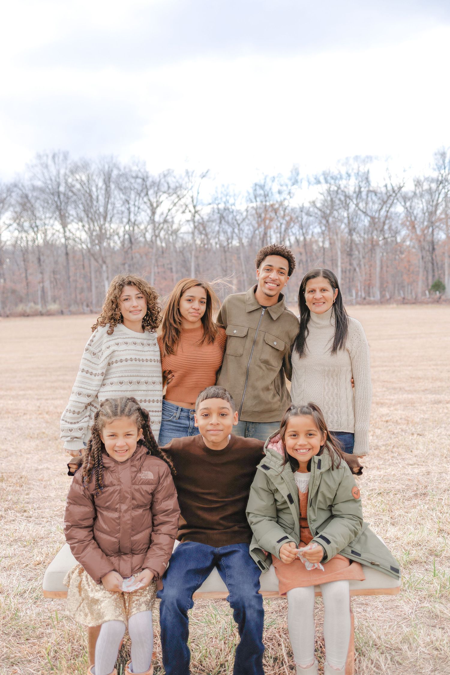 Group of seven children posing outdoors in a field during fall, wearing cozy sweaters and jackets for a family portrait.