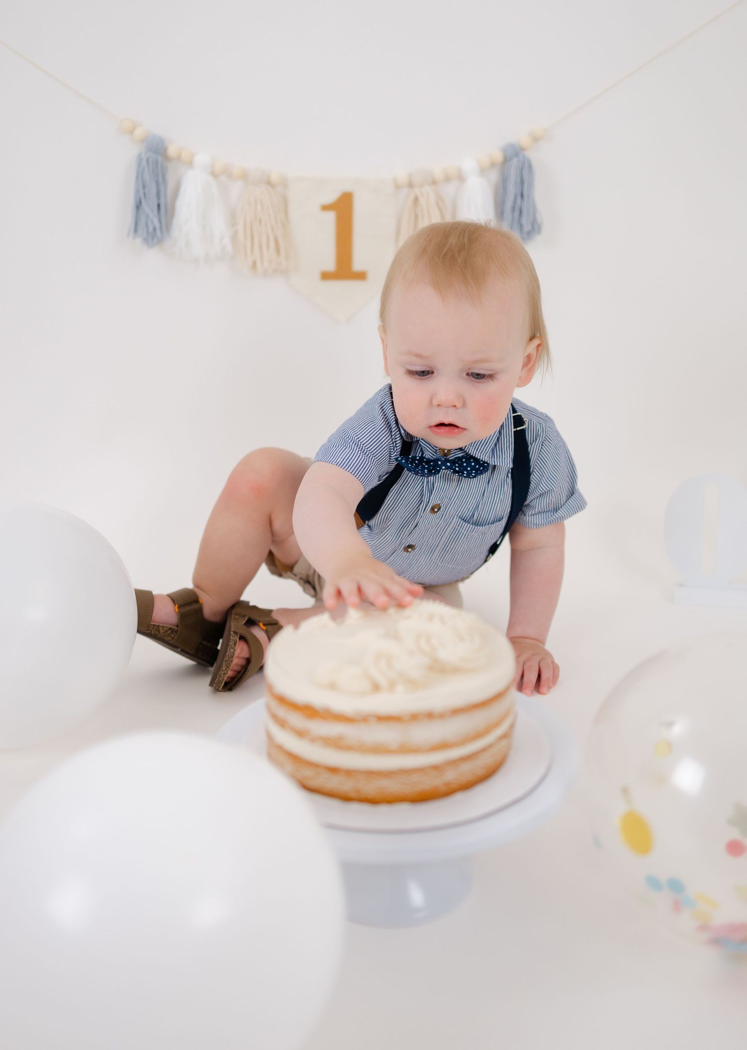 Baby boy in suspenders touches his first birthday cake during a cake smash photo session with balloons.