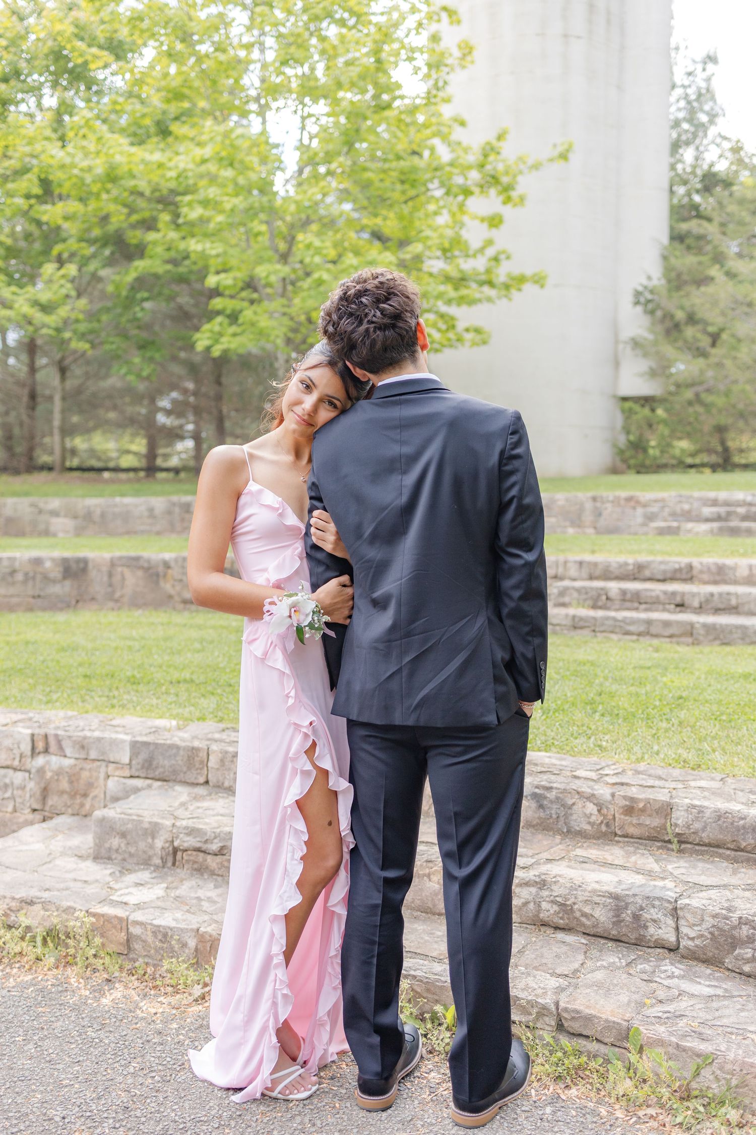 A young couple in prom attire, girl in pink gown with corsage, boy in dark suit, posing outdoors near stone steps.