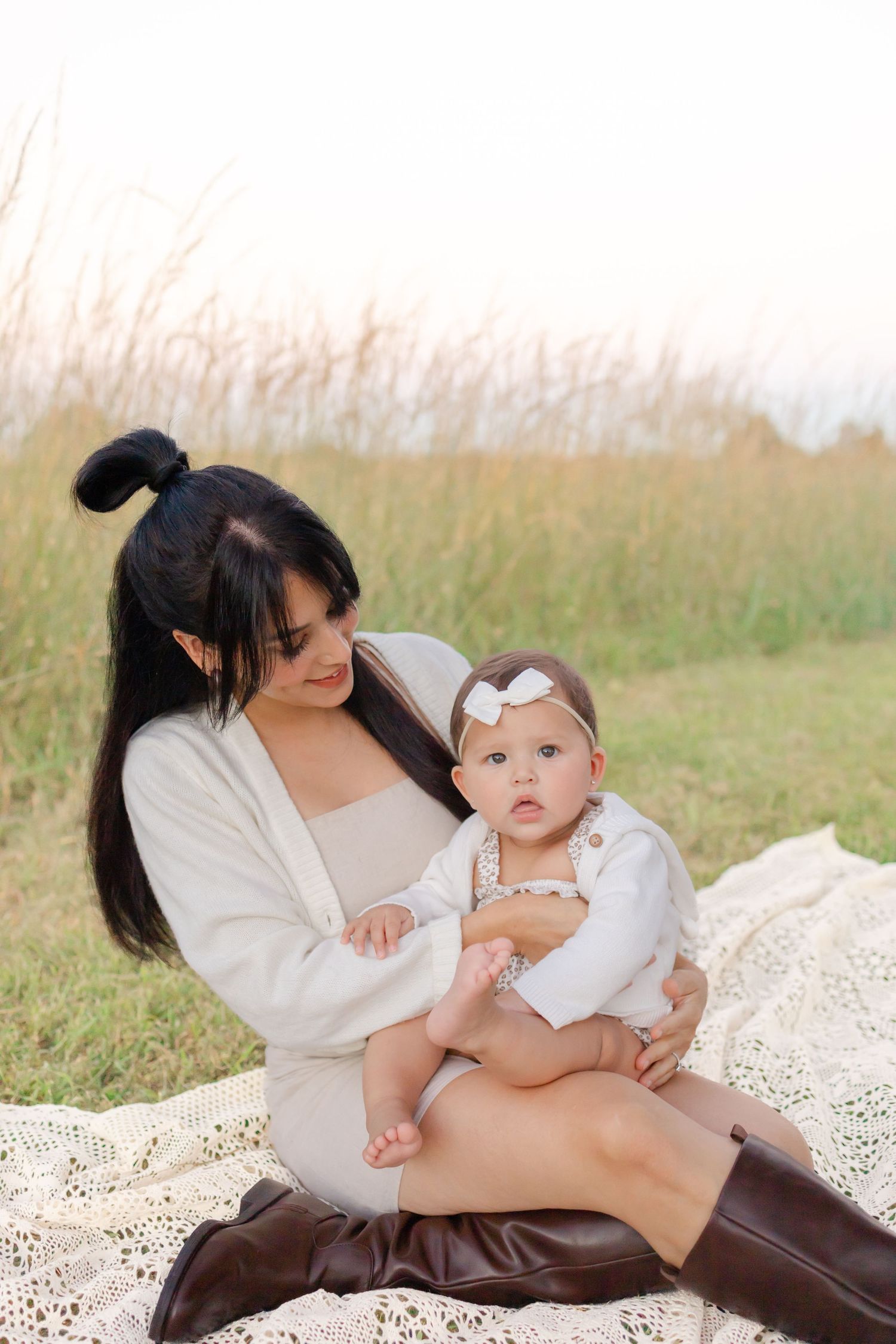 A mother with long dark hair holds her baby girl in a white bow headband outdoors on a lace blanket.