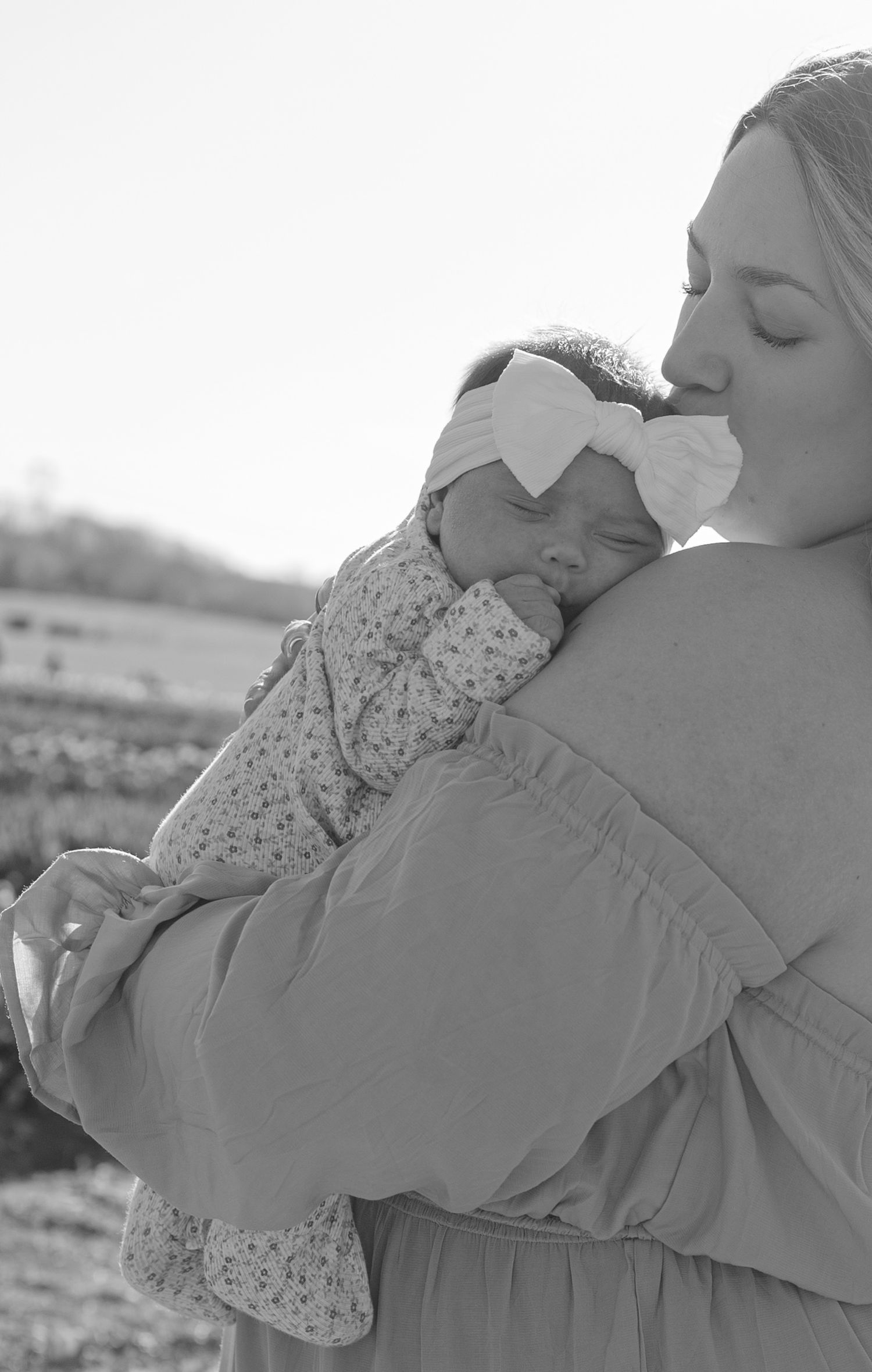 Black and white photo of a mother tenderly kissing her newborn baby wrapped in a floral outfit with a bow headband.