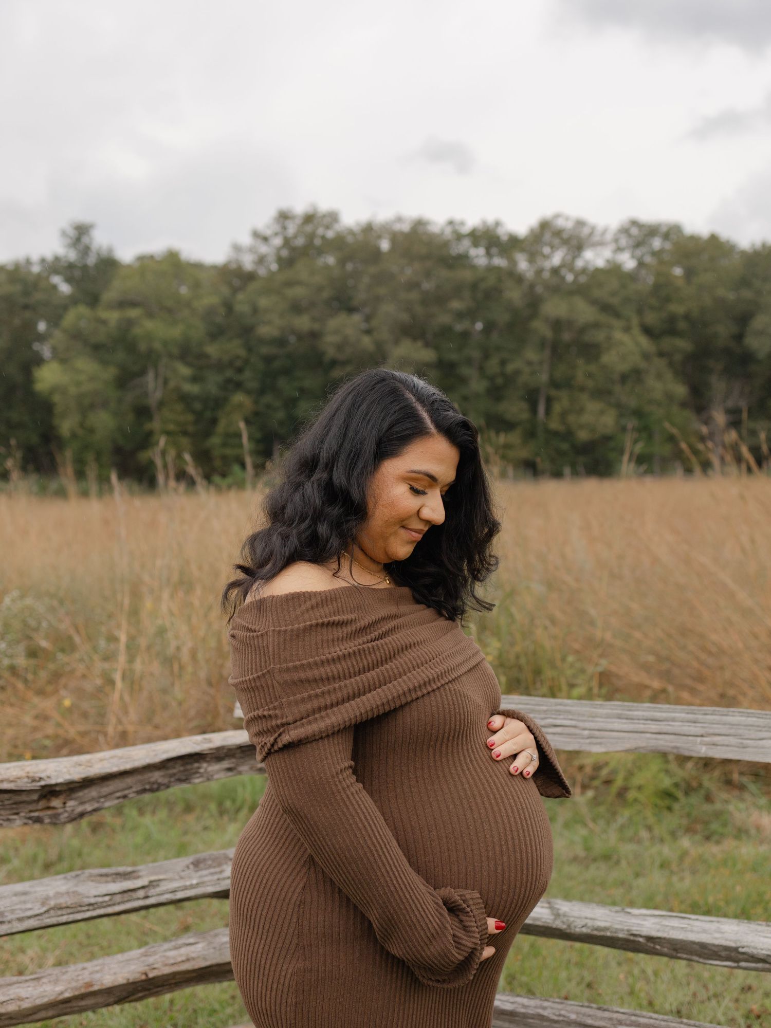 Pregnant woman in brown off-shoulder ribbed dress cradling baby bump outdoors near rustic fence and golden field.