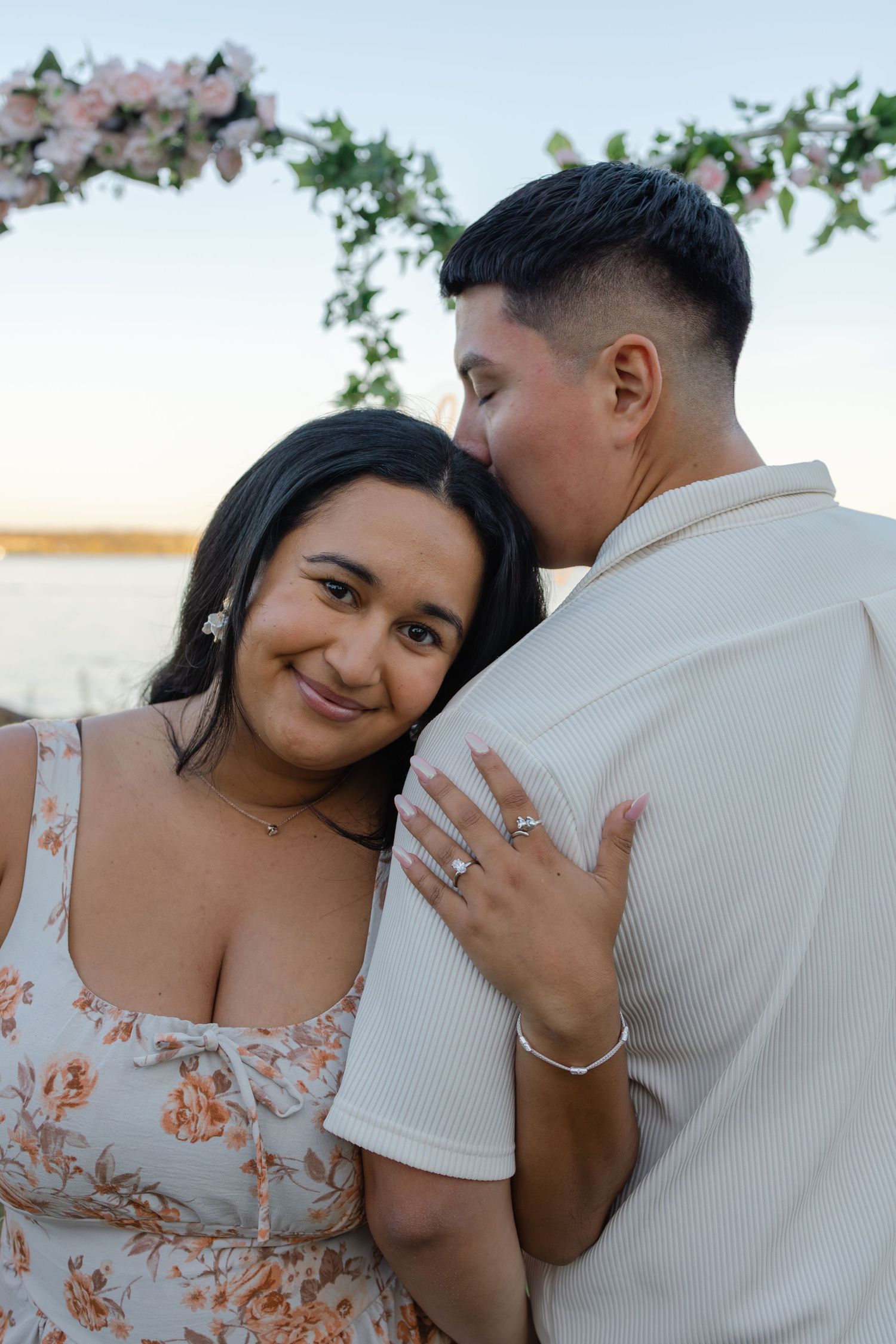 Couple sharing a romantic moment at sunset, woman in floral dress smiling with engagement ring, man kissing her cheek near floral arch.