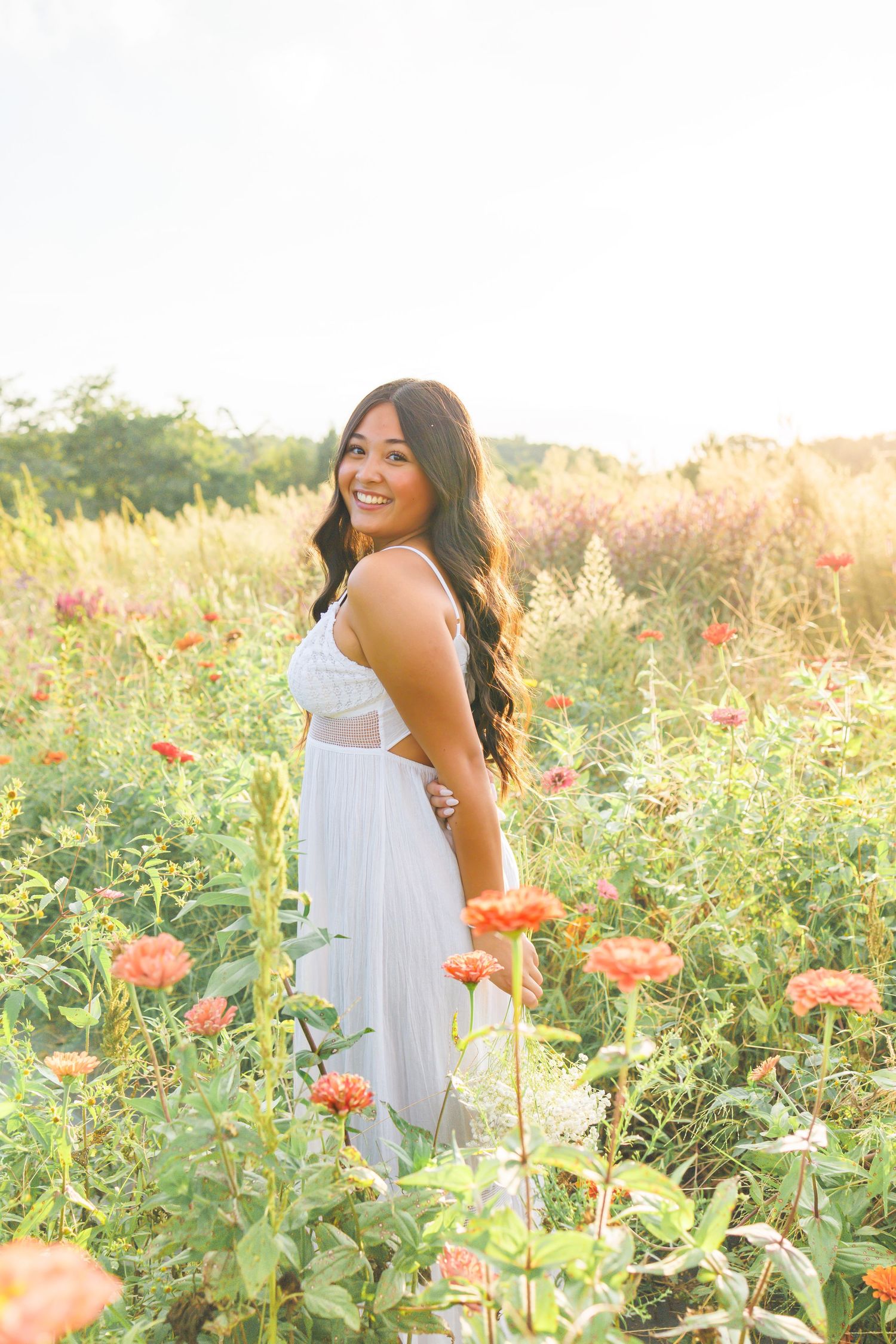 Smiling woman in white dress standing among vibrant orange zinnia flowers in a sunlit meadow field.