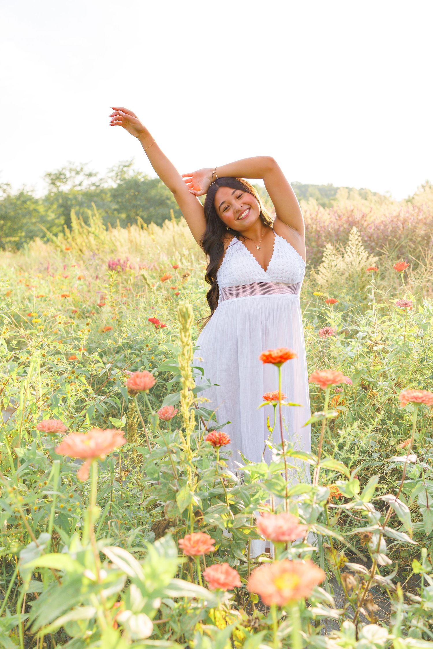 Smiling woman in white dress posing with arms raised in a vibrant field of orange flowers on a sunny day.
