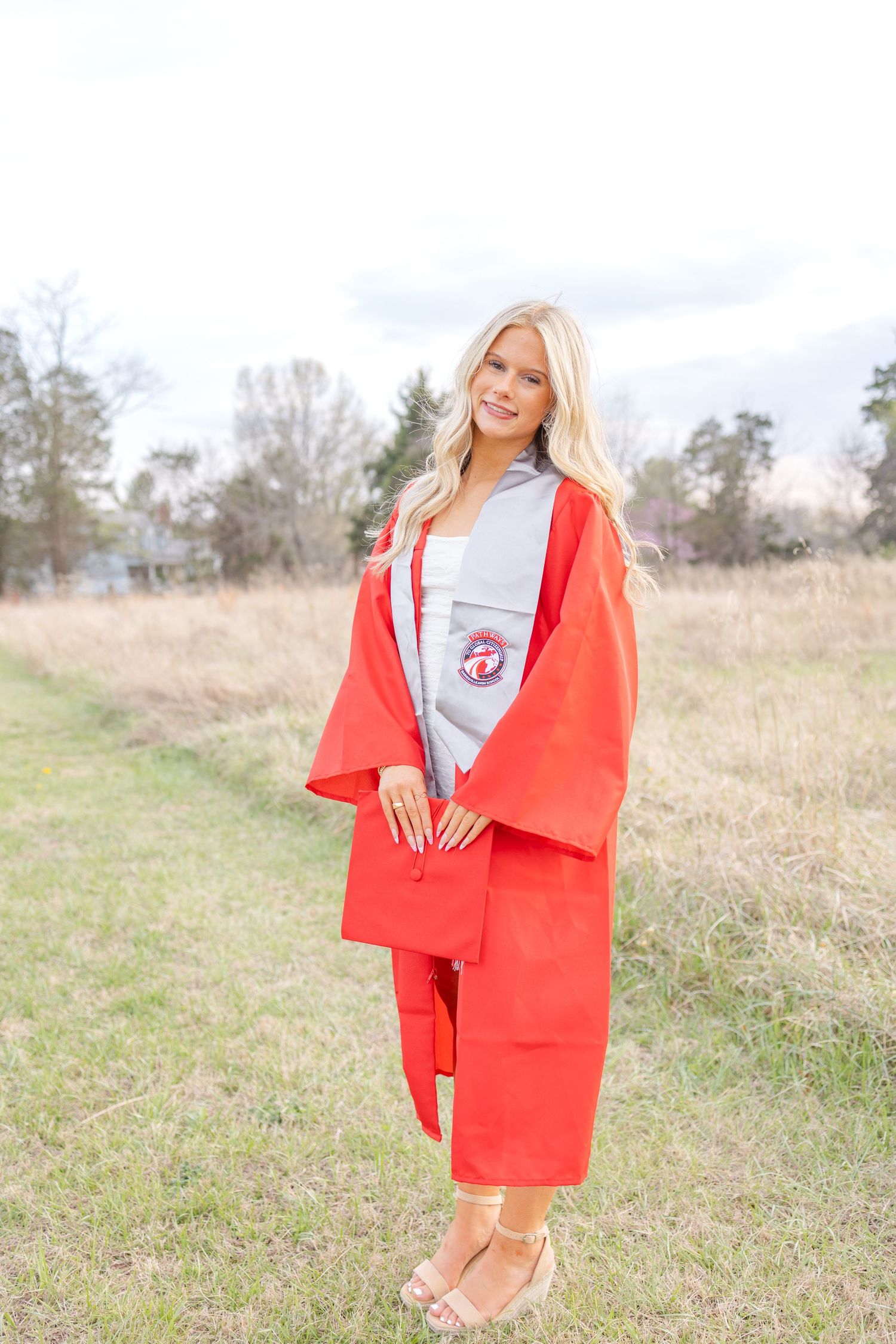 Smiling blonde graduate in red cap and gown standing outdoors in a field on a cloudy day.