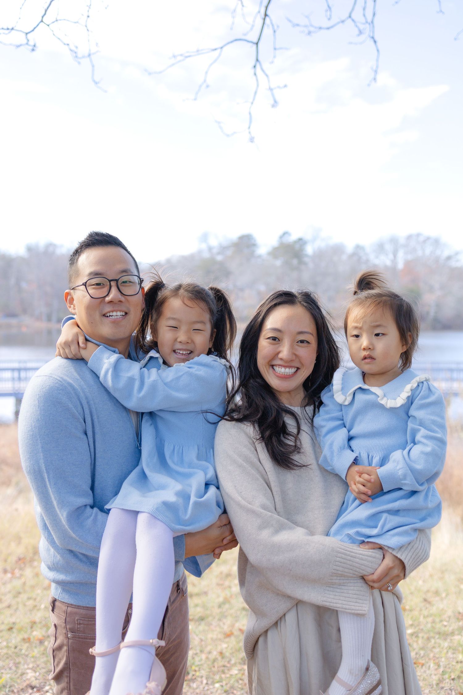 A smiling Asian family of four poses outdoors near a lake, parents holding two young daughters in matching blue outfits.