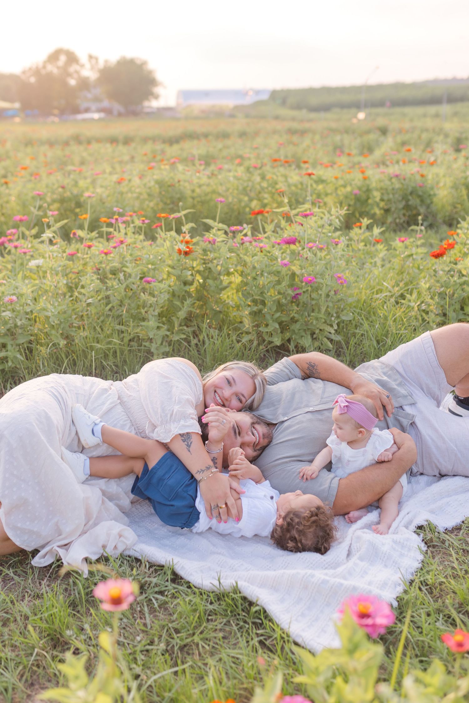 Family of five lying on a blanket in a colorful flower field at sunset, smiling and cuddling together.