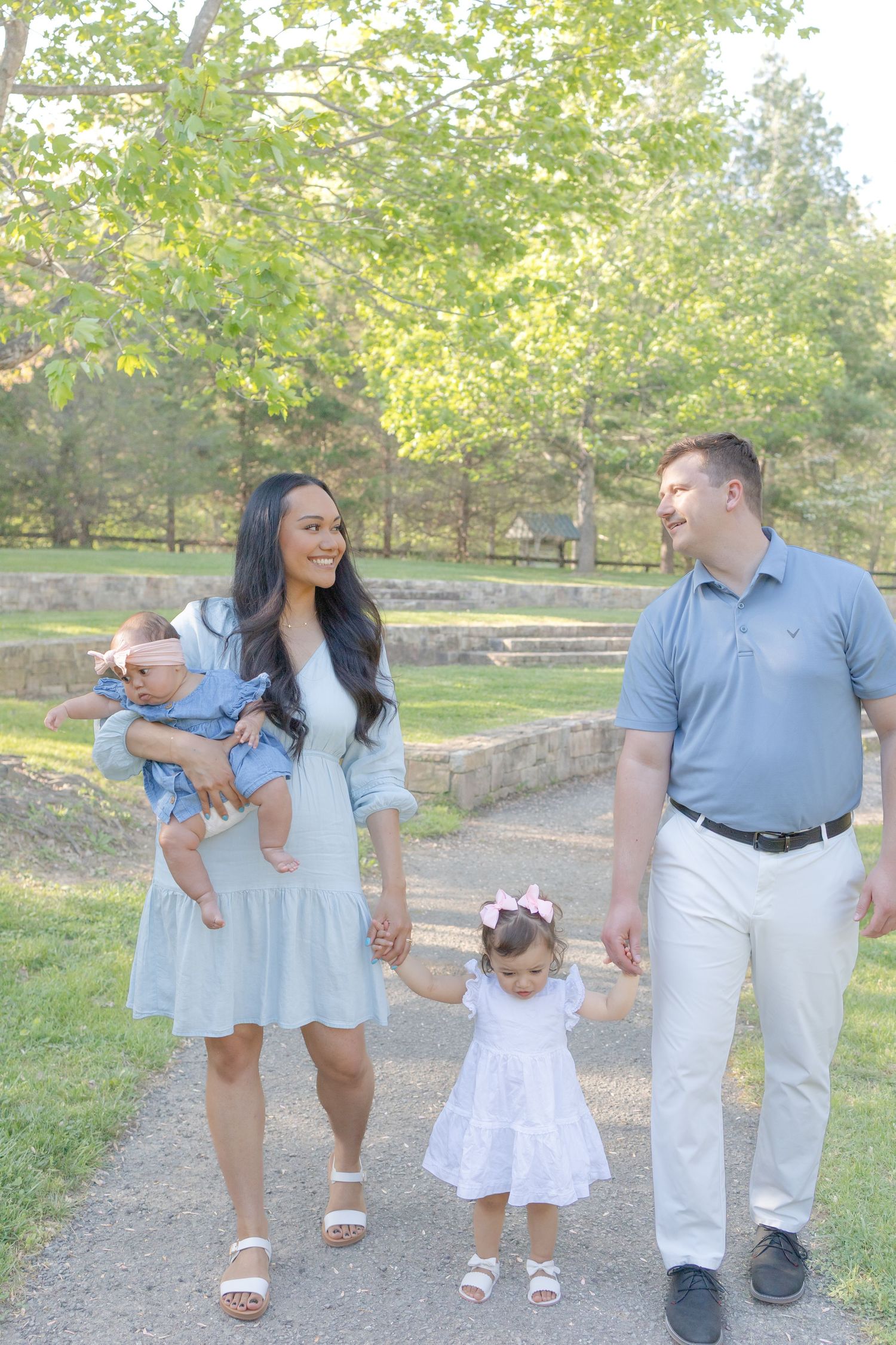 Happy family of four walking on a park path, parents holding hands with toddler girl in white dress.