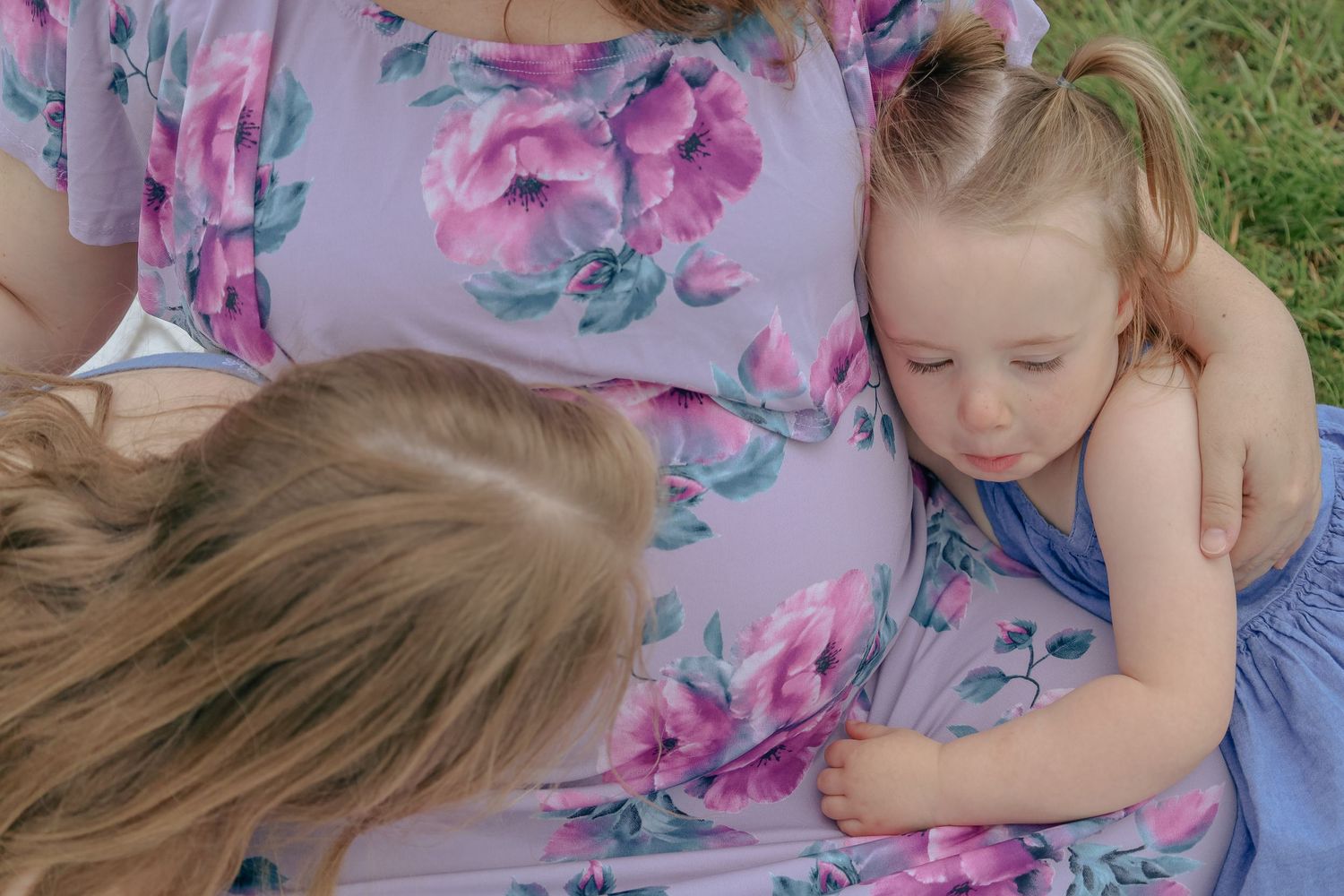 Pregnant woman in floral dress with two young children resting against her baby bump outdoors.