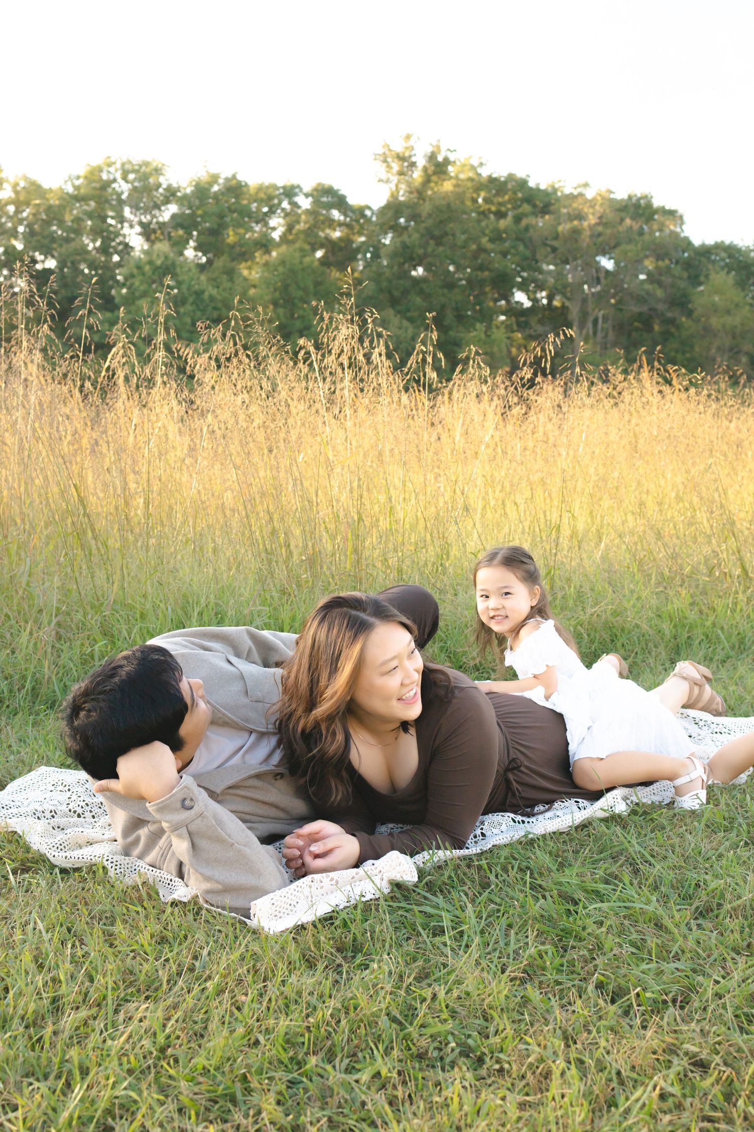Family enjoying a sunny outdoor picnic on a blanket in a grassy meadow, with two adults and a young child smiling together.