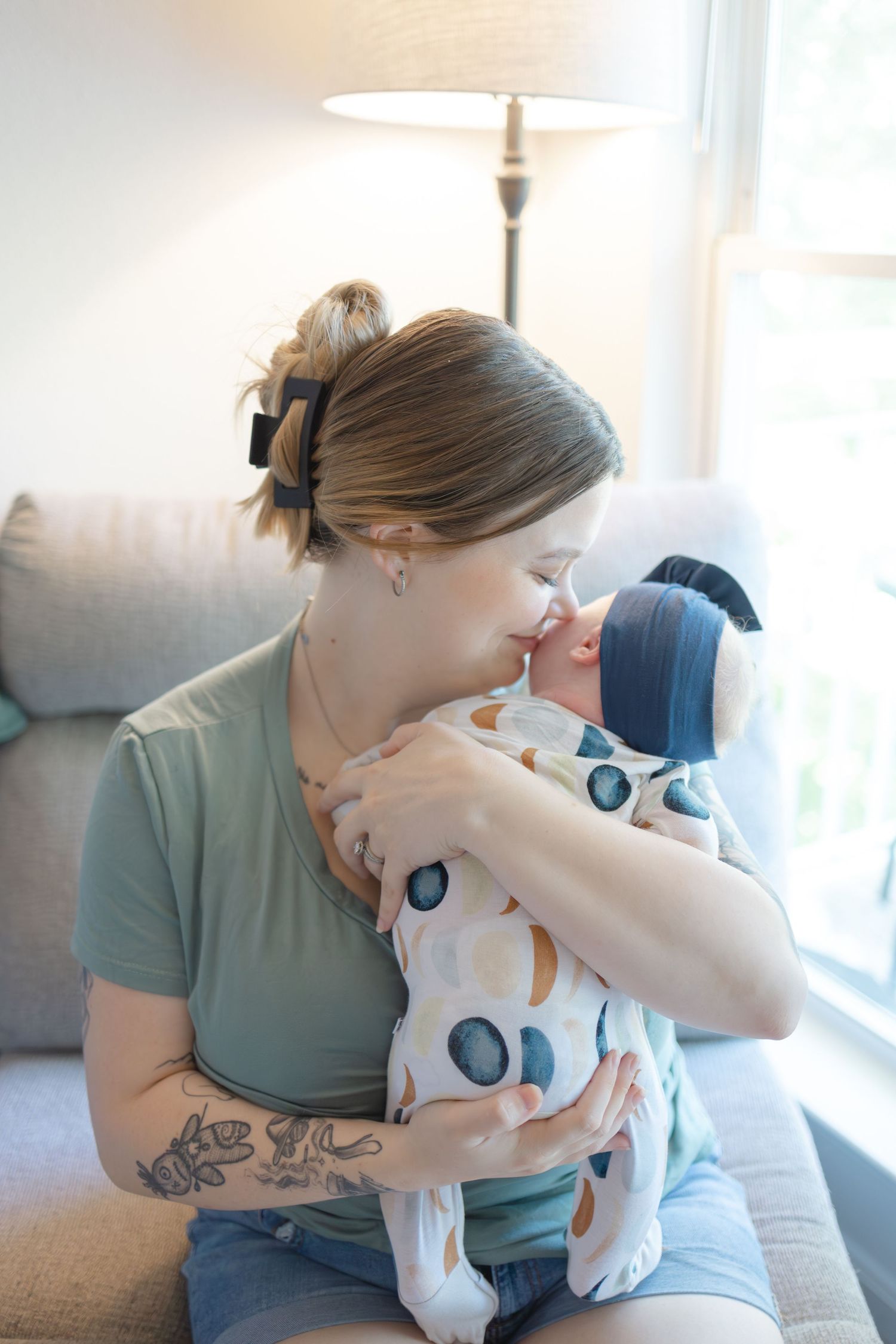 Parent in teal shirt holds and comforts crying baby in white onesie on couch in sunlit living room.