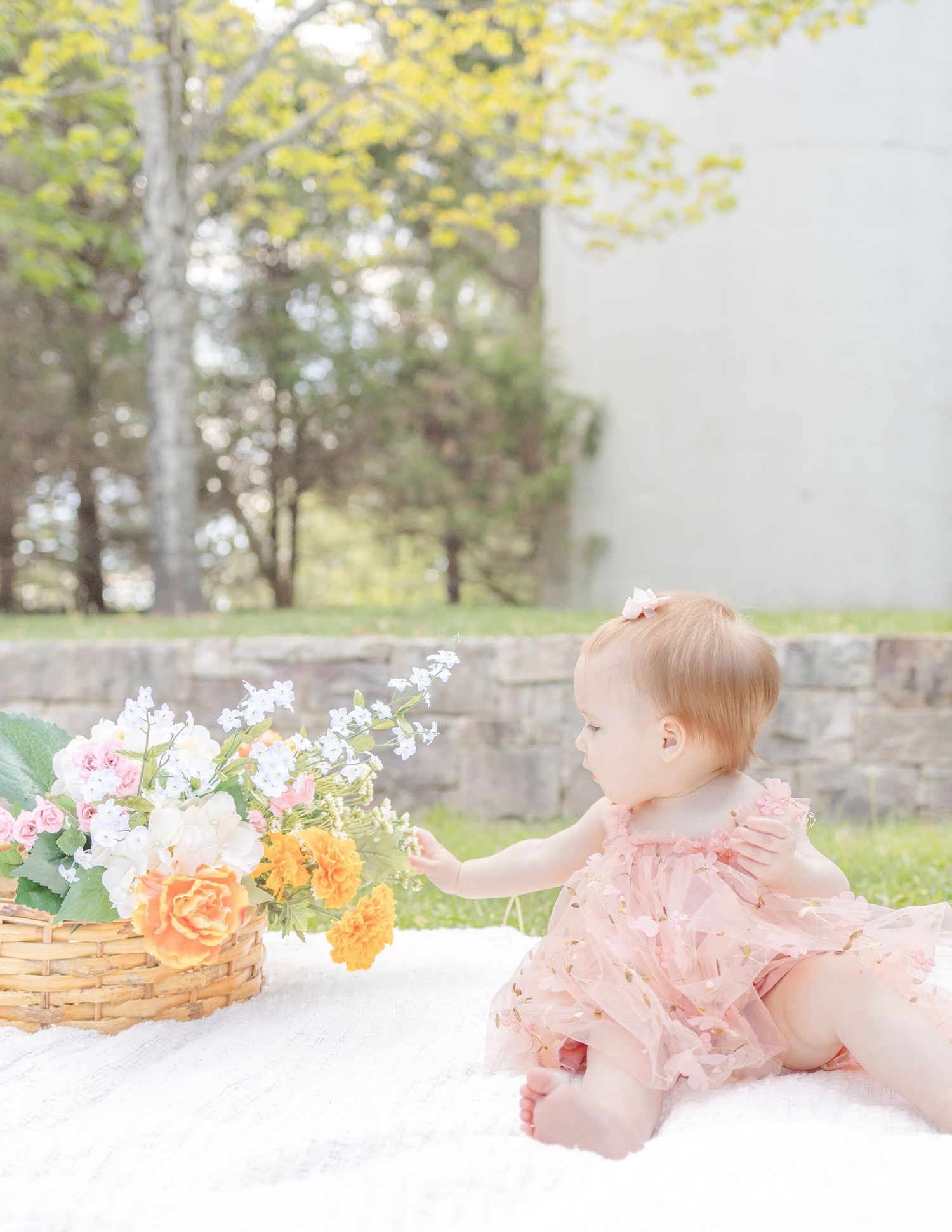 Baby girl in pink dress reaching for flowers in a wicker basket on a white blanket outdoors.