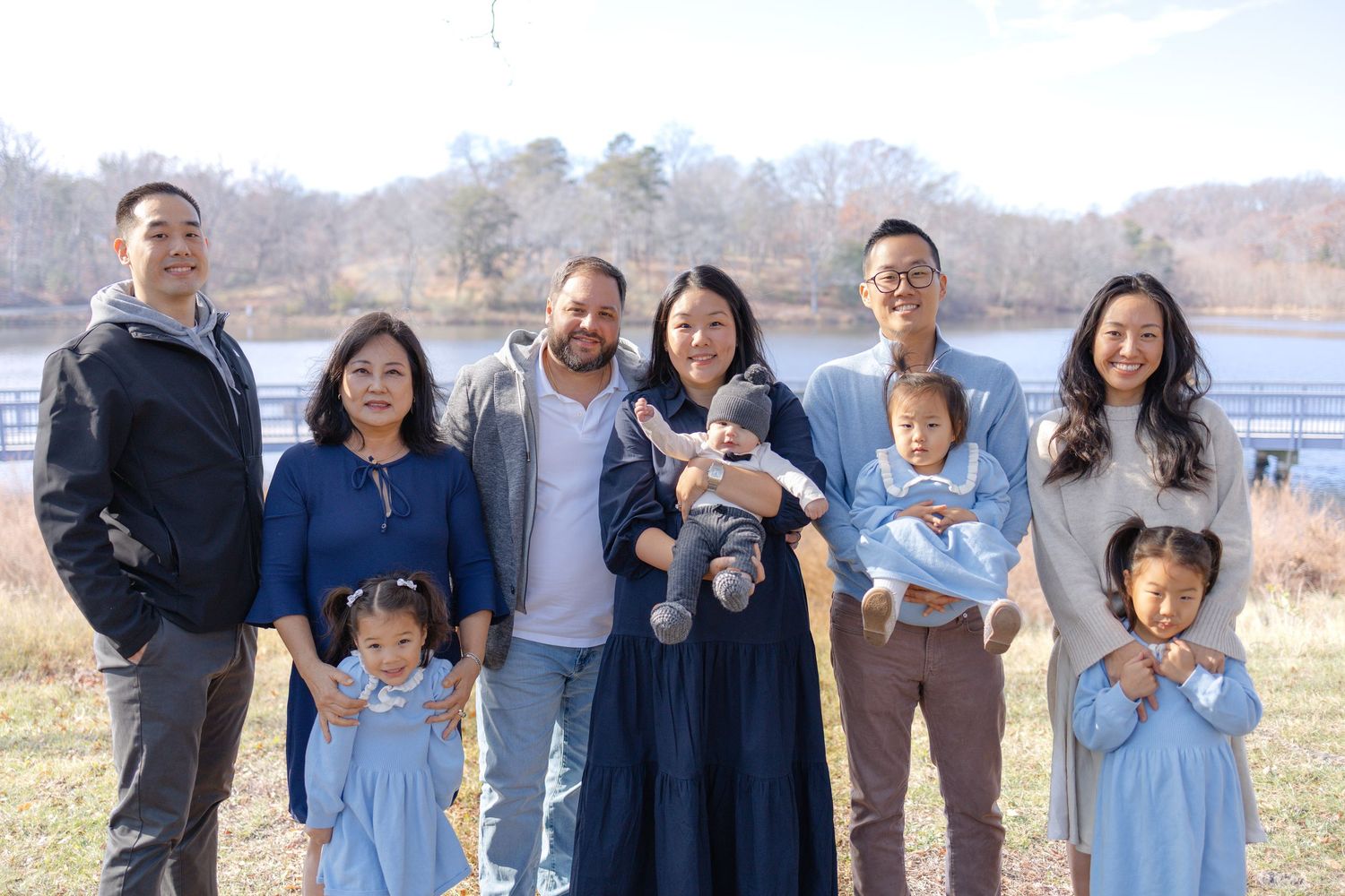 A joyful multigenerational Asian family poses outdoors near a lake on a sunny winter day, dressed in coordinating blue outfits.