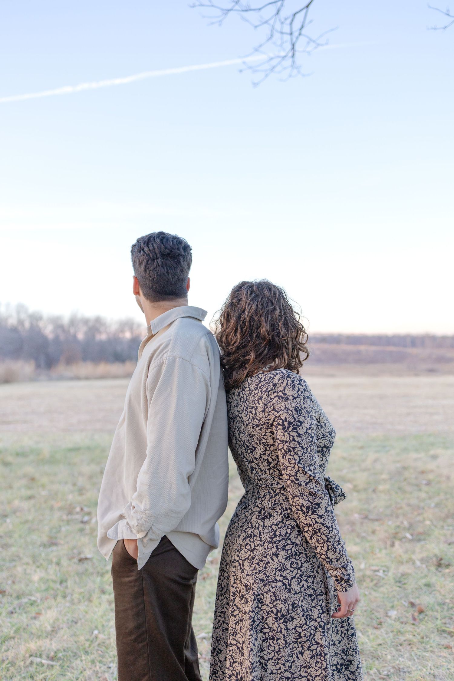 Couple standing back-to-back in an open field during golden hour, wearing casual and floral dress attire.