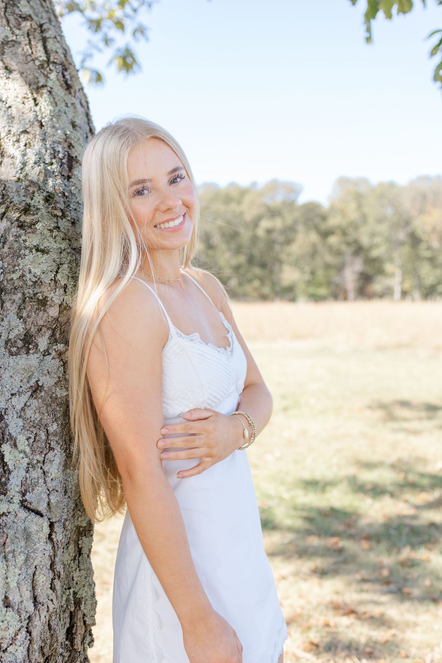 Woman in white sundress poses by tree trunk in sunny outdoor setting with natural field background.