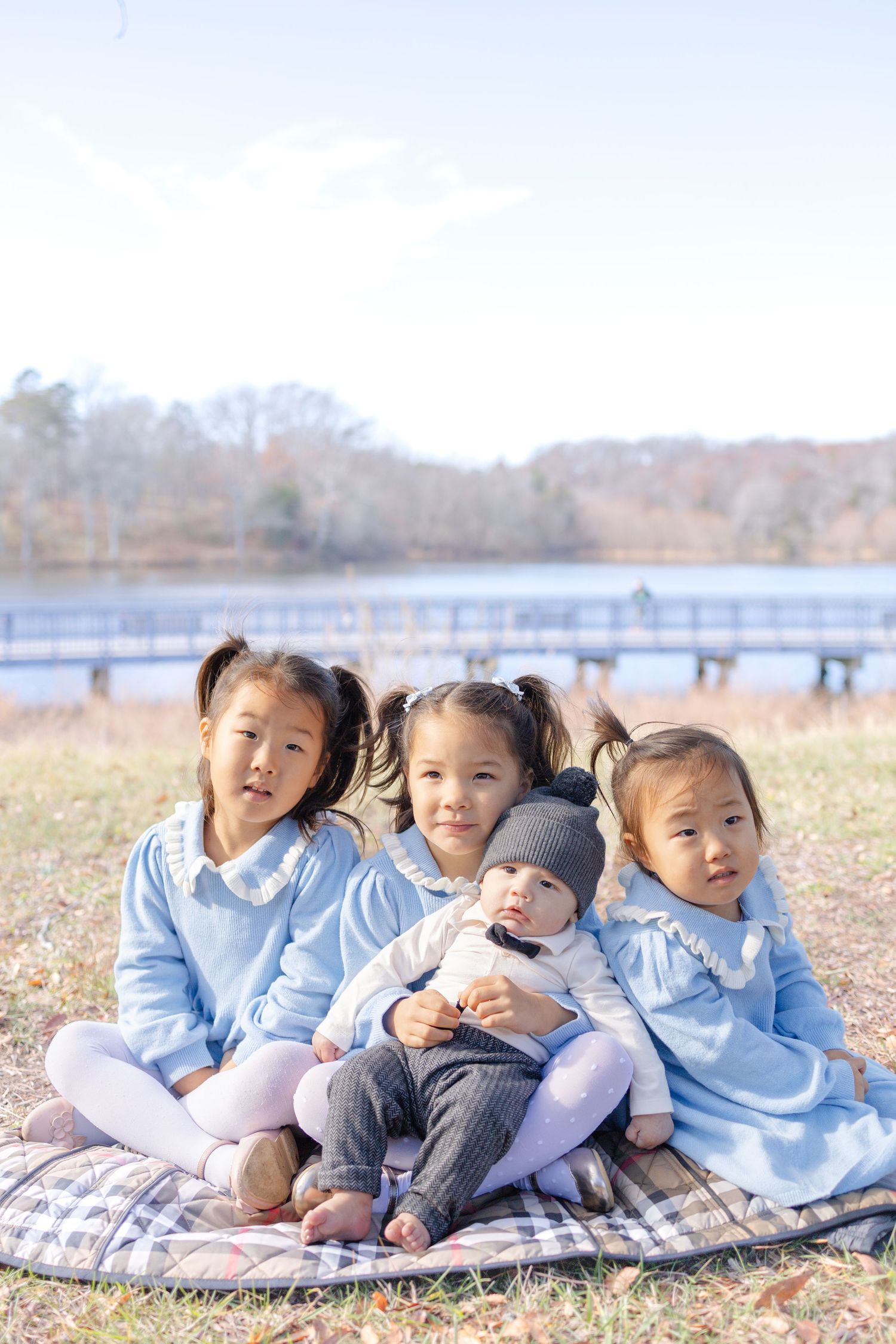 Four young children in matching blue outfits sit on a blanket outdoors near a lake on a sunny day.