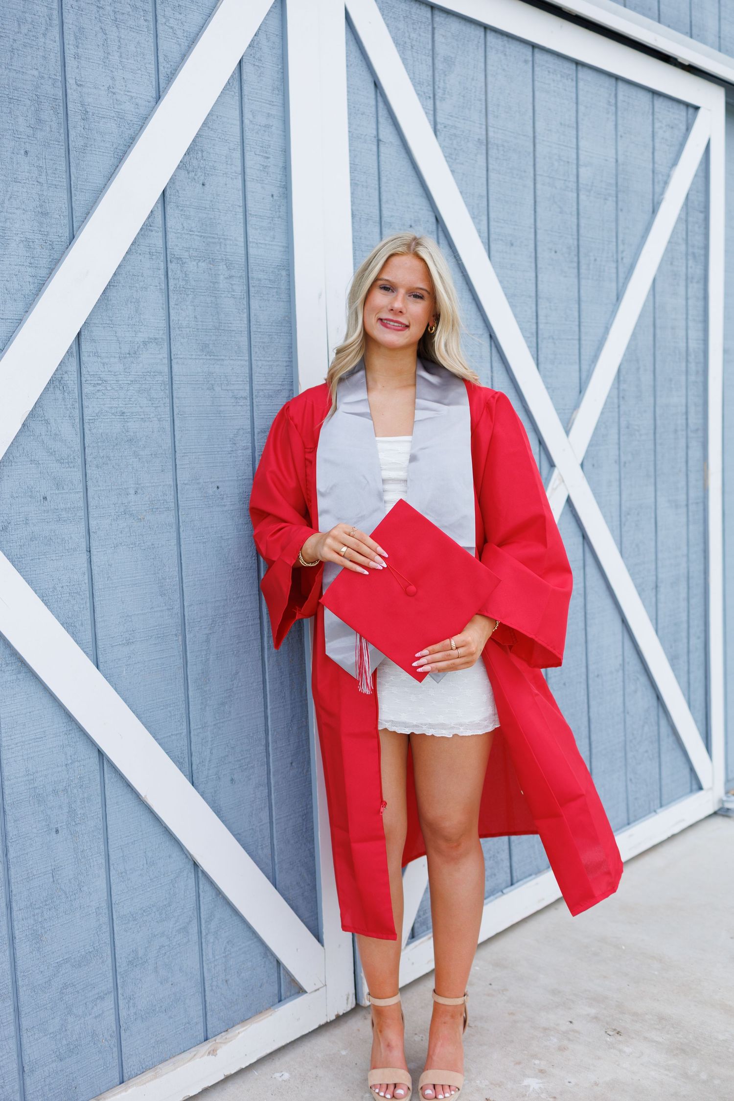 Smiling blonde woman in red graduation cap and gown holding diploma, posing against blue barn door.