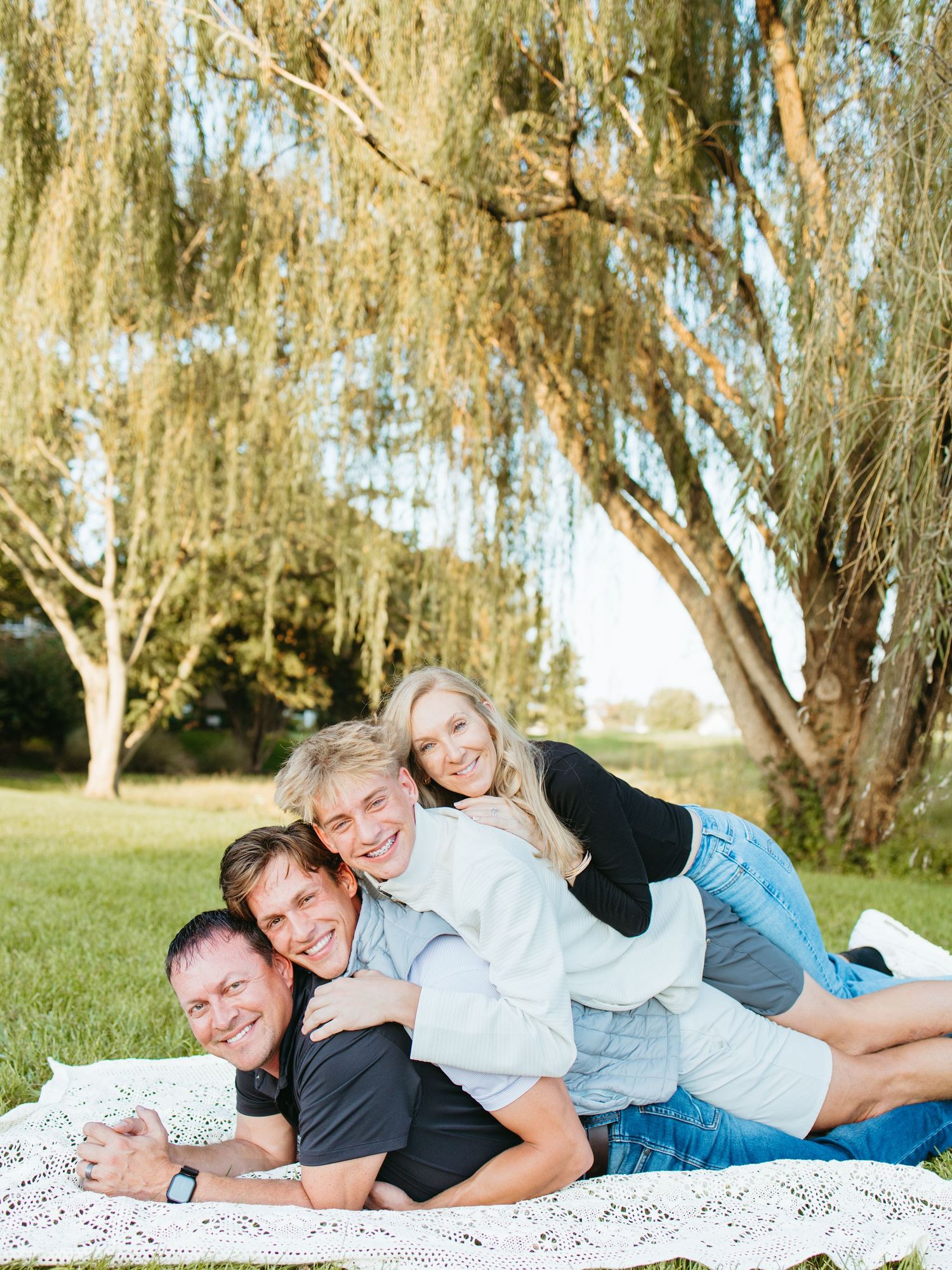 Family of four laughing and stacking on a blanket under a weeping willow tree in a green park.