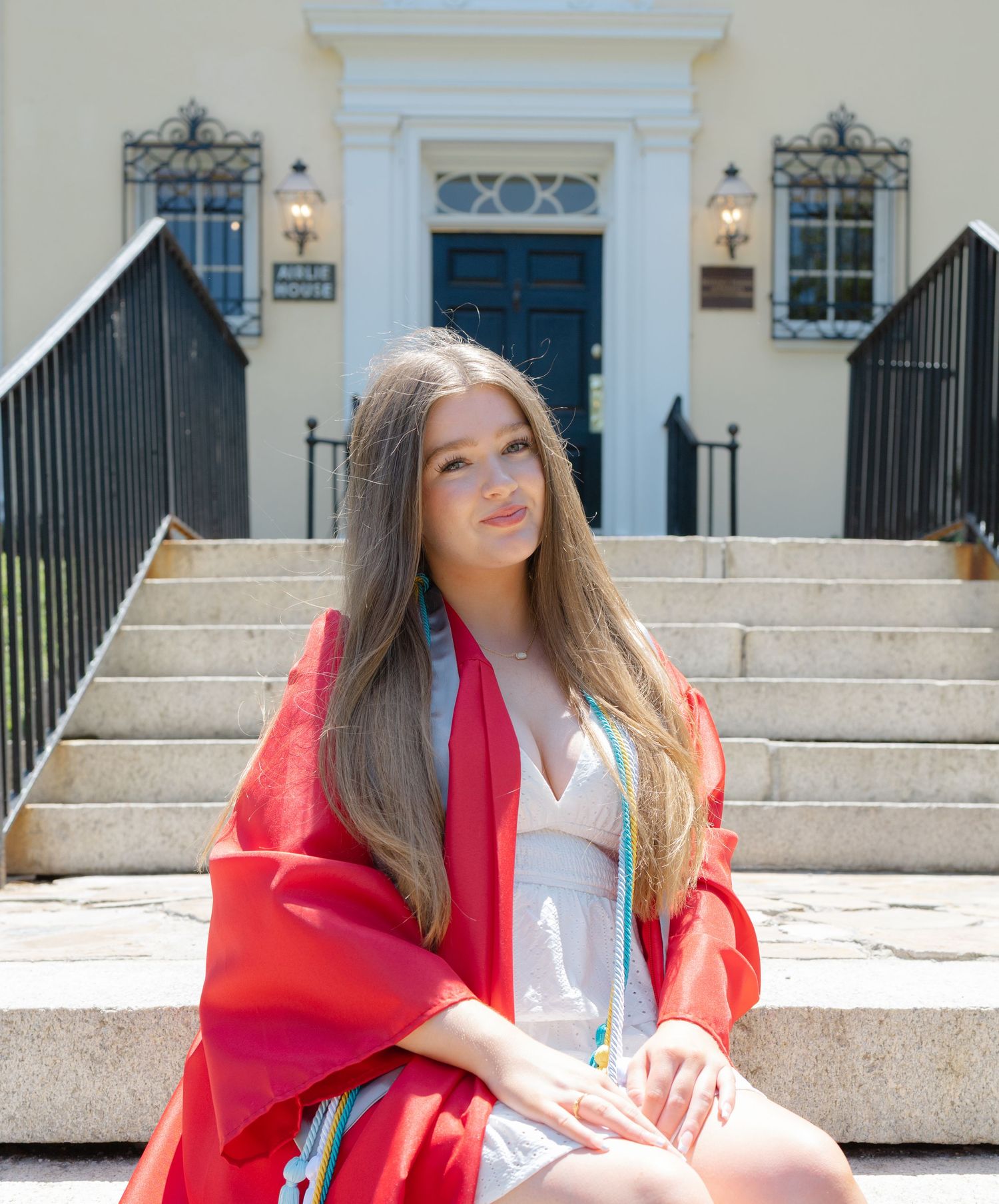 Graduate in red regalia sits on steps of white building holding diploma.
