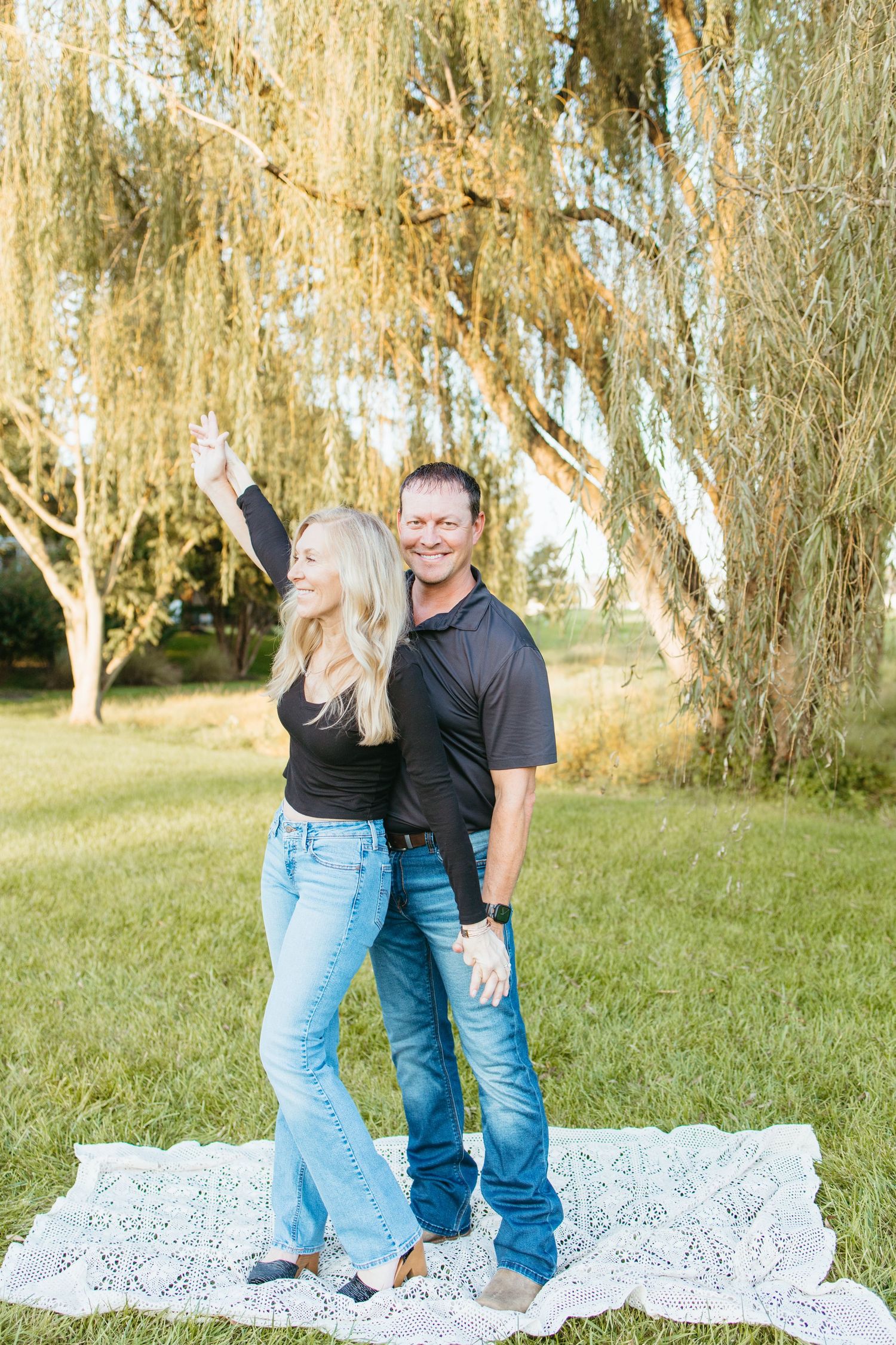 A smiling couple in jeans and black tops stands on a lace blanket under a weeping willow tree in a sunny park.