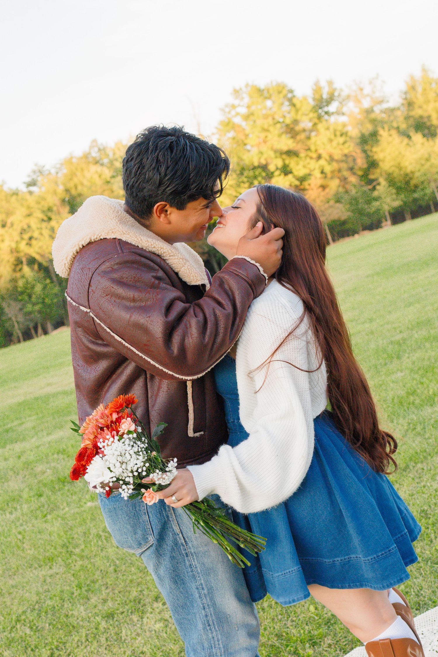 Couple sharing a romantic kiss outdoors, man holding red and white flowers in a green park setting.