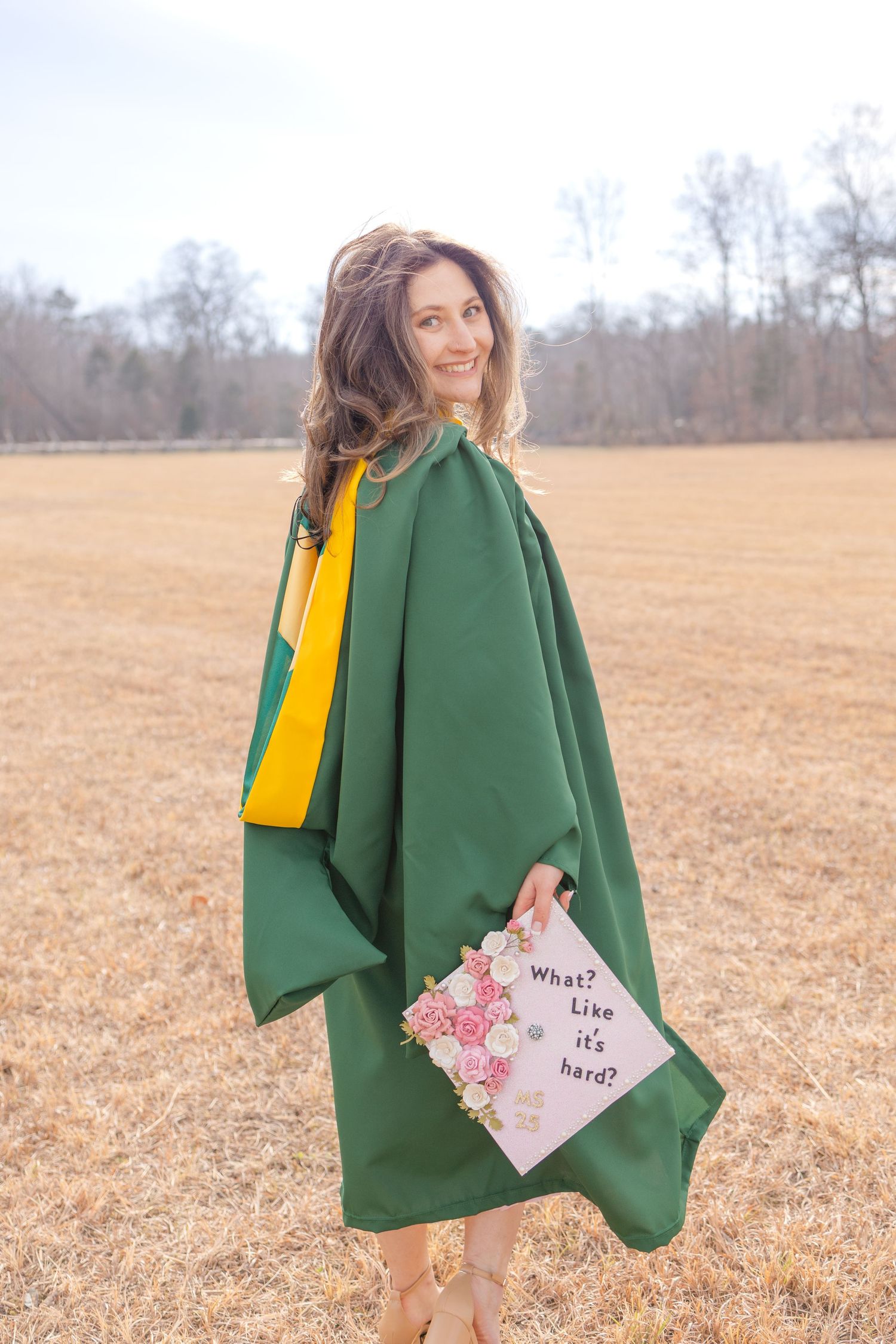 Young woman in green graduation gown holding decorated cap reading 'What? Like it's hard?' in an open field.