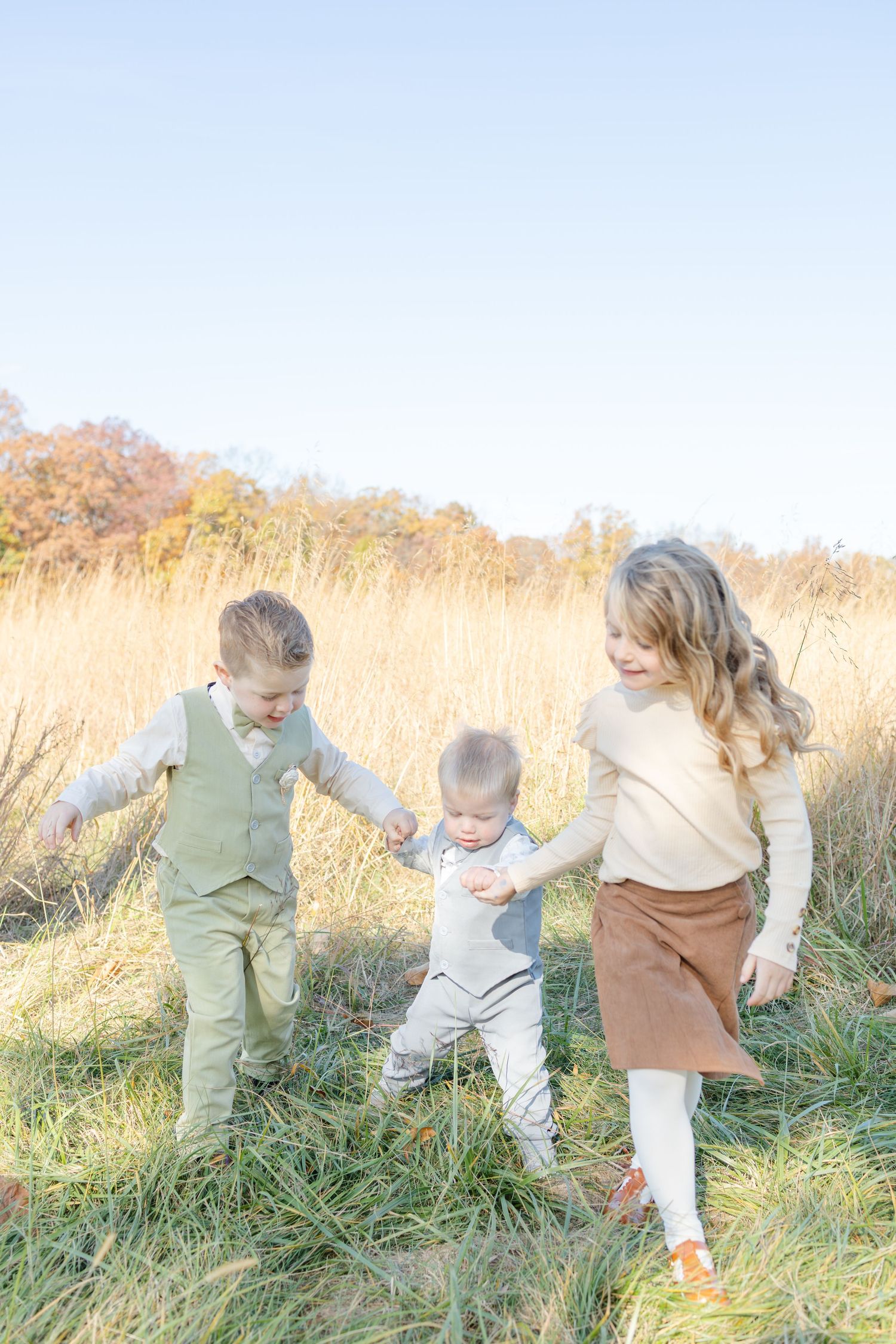 Three young children playing together in a golden autumn field, with the oldest boy and girl helping a toddler walk.