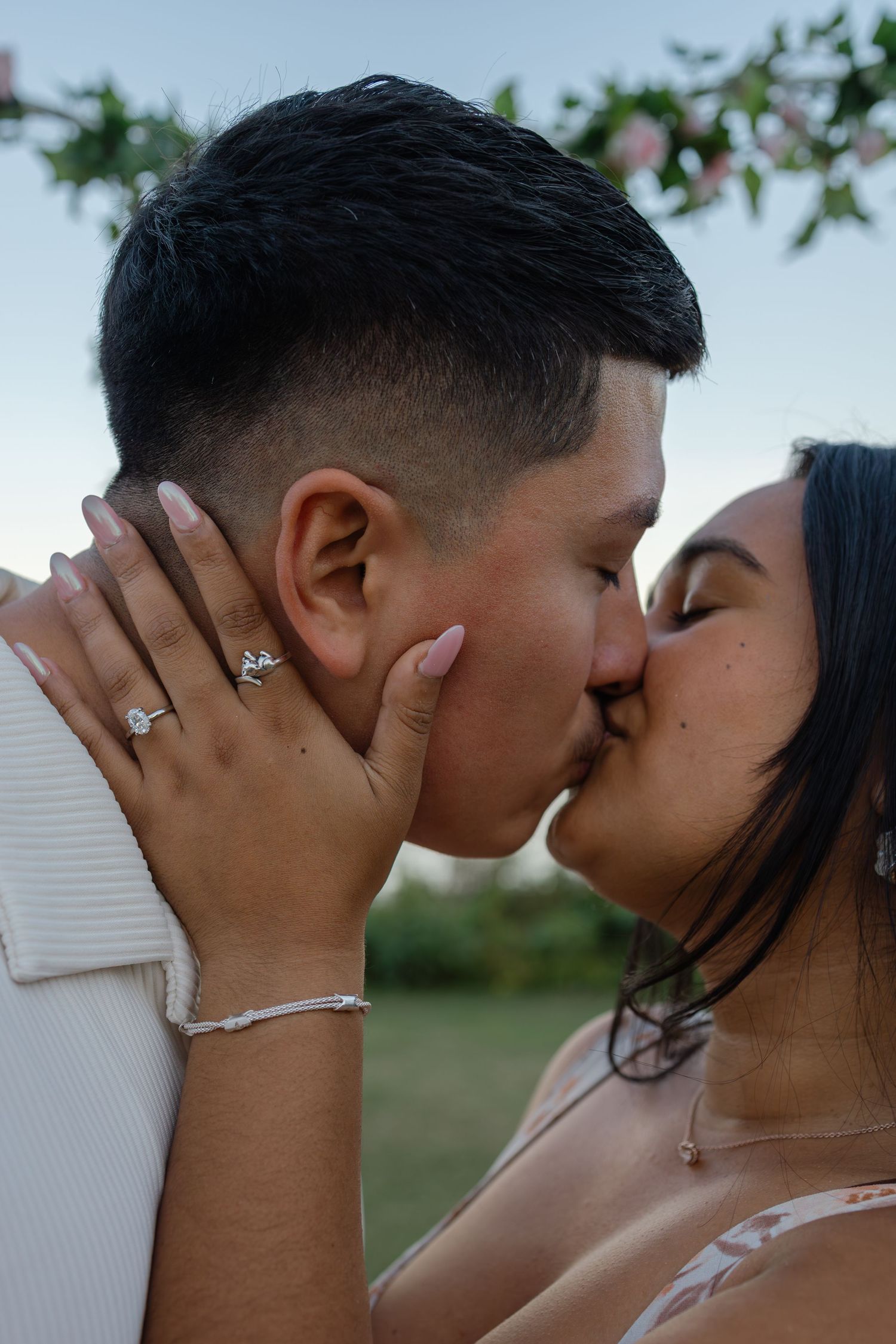A romantic couple shares a tender kiss outdoors with greenery in the background.