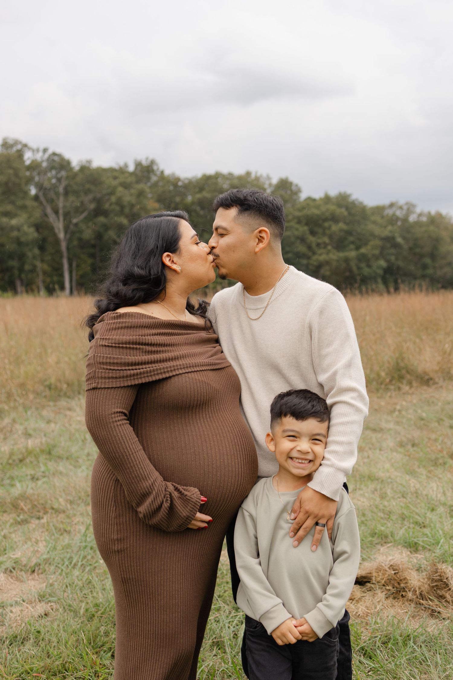 Pregnant woman in brown dress kisses partner while smiling boy stands between them in a field.