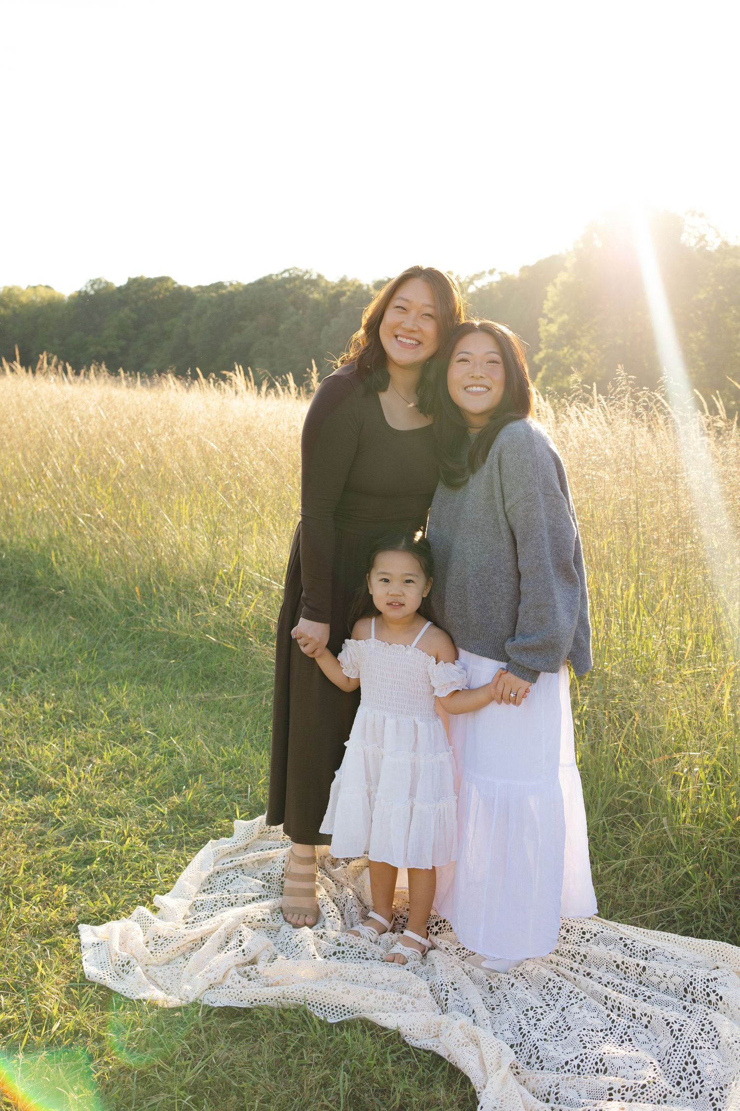 Three smiling women and a young girl in white dress pose in a sunny golden field during a family portrait session.