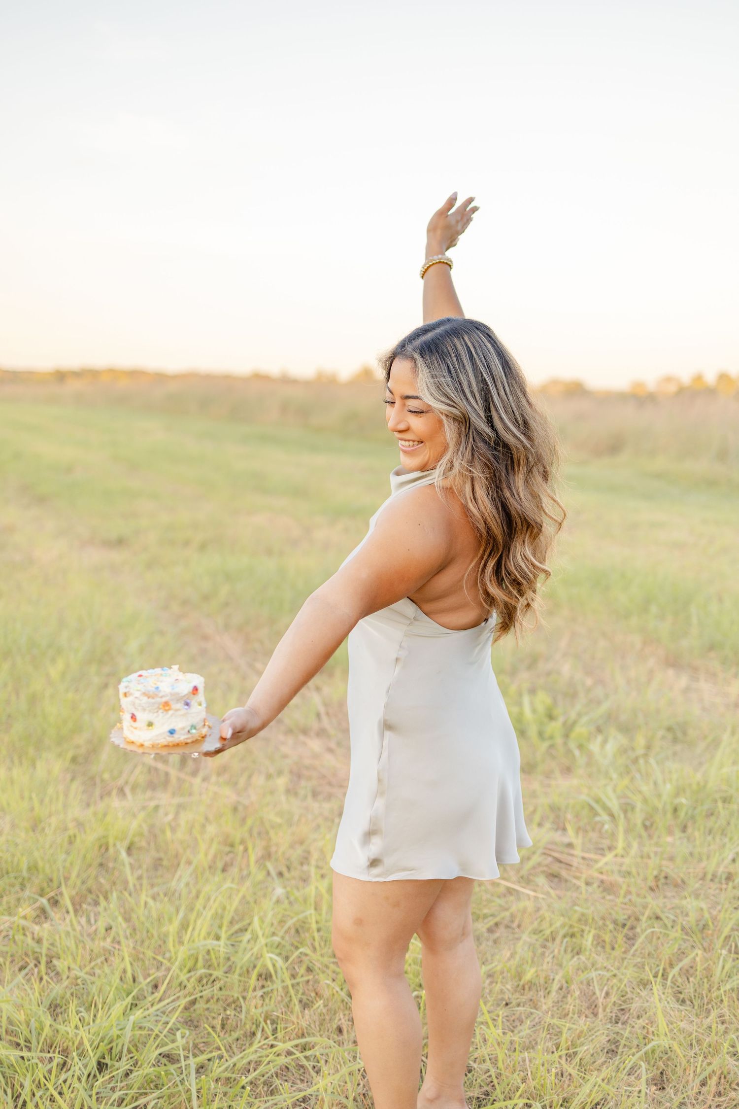 Woman in sage mini dress holding a birthday cake in a golden field at sunset, hair blowing in the breeze.