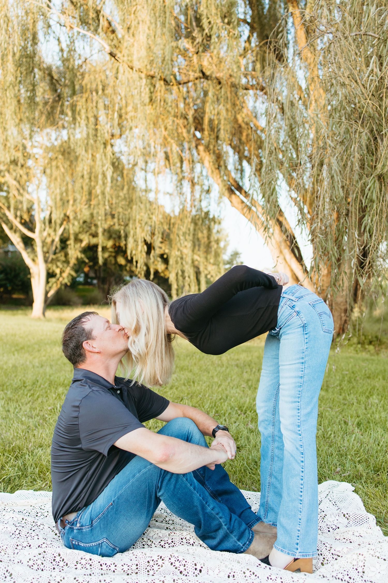 Couple sharing a romantic moment outdoors on a blanket under a willow tree, man seated kissing woman leaning down.